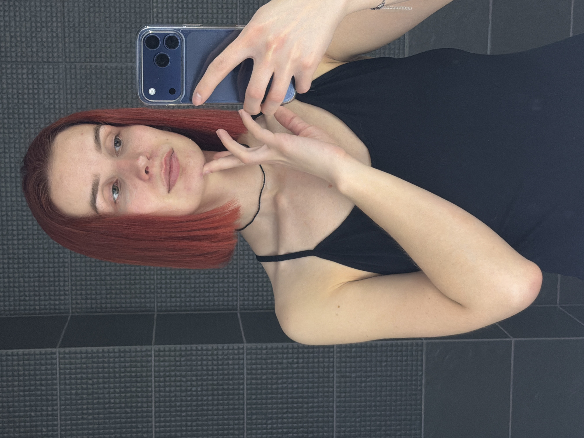 A fair-skinned woman with dyed red hair takes a selfie in what appears to be a bathroom or tiled room. She is holding a blue smartphone, likely an iPhone, with multiple cameras visible on the back. She is wearing a black, spaghetti-strap top. The background features a wall or floor covered in small, textured tiles. There is no information available in the image to suggest that the location is in the city of Chaikovsky, Russia.