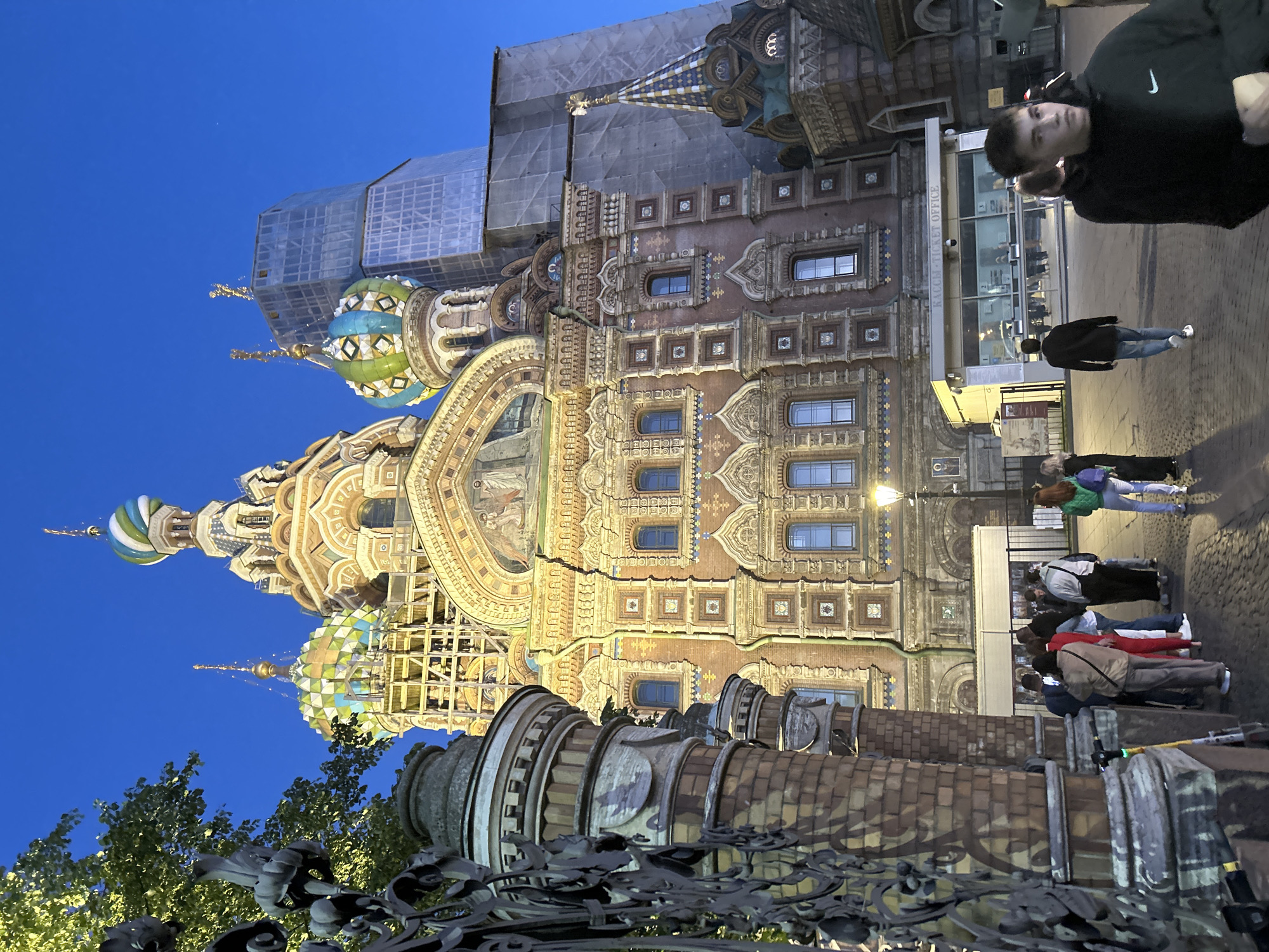This is a full-frame, eye-level, vertical outdoor shot of the Church of the Savior on Spilled Blood in Saint Petersburg, Russia, during twilight. The church's intricate, colorful onion domes are prominently featured against a deep blue sky. To the right of the church, a modern skyscraper is visible, partially covered in scaffolding and construction netting. In the foreground, a decorative wrought-iron fence and ornate brick pillars frame the scene on the left. A group of people are gathered near a ticket office building in front of the church. One young man in the foreground is walking away from the camera towards the right. The ground is paved with cobblestones.