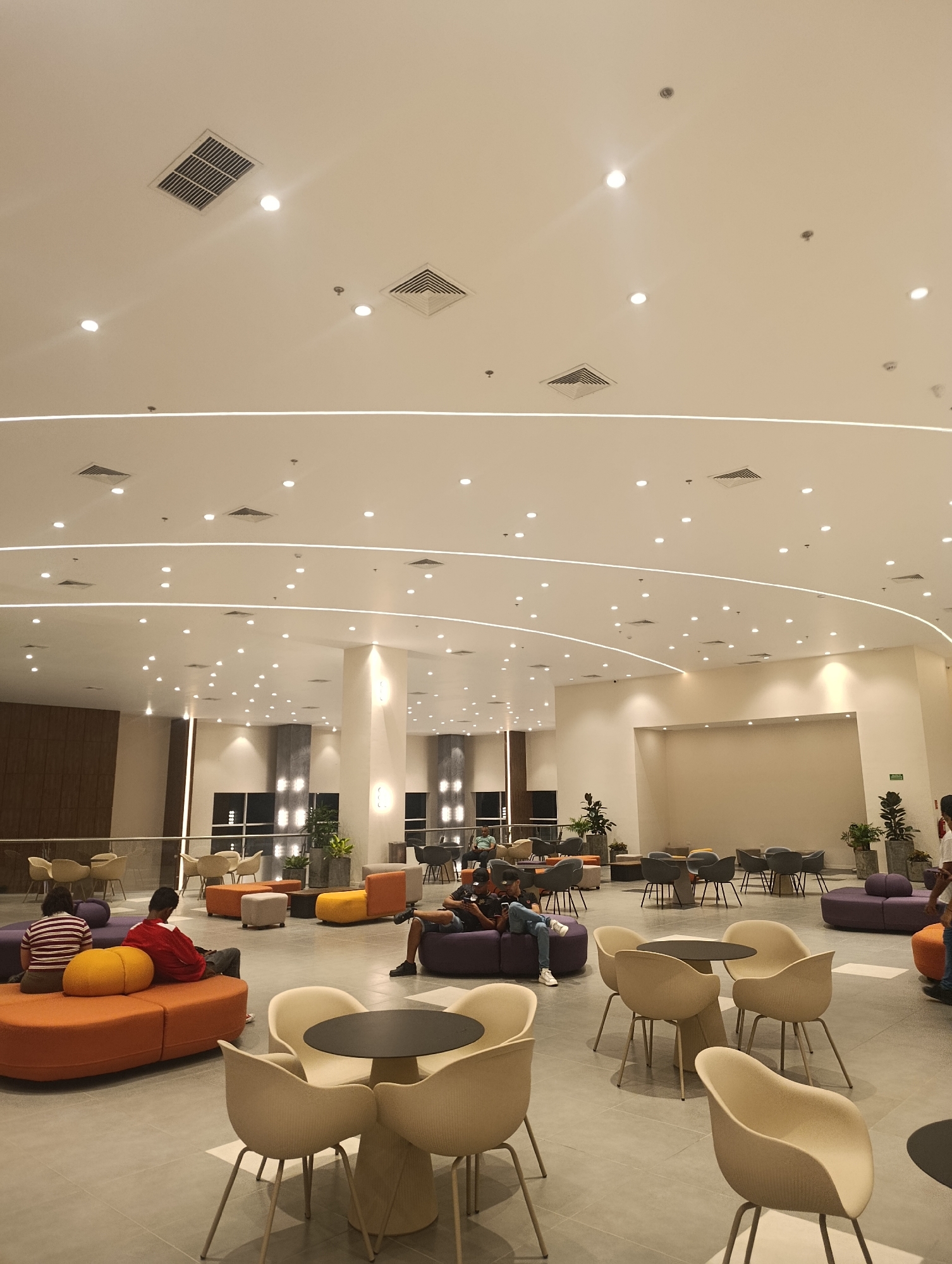 The image depicts a spacious, brightly lit indoor lounge area, likely in a commercial or public building in Porlamar, Venezuela. The room features a high ceiling with multiple recessed lights and decorative curved linear lighting. The floor is tiled in a neutral color.
The lounge is furnished with a variety of seating arrangements, including modular orange and purple couches, individual chairs in neutral tones paired with small round tables, and potted plants are scattered throughout the area. Several people are present, some sitting and relaxing on the couches while others are seated at the tables. They appear to be engaged in casual conversation and individual activities like using mobile phones. The overall atmosphere is relaxed and inviting.