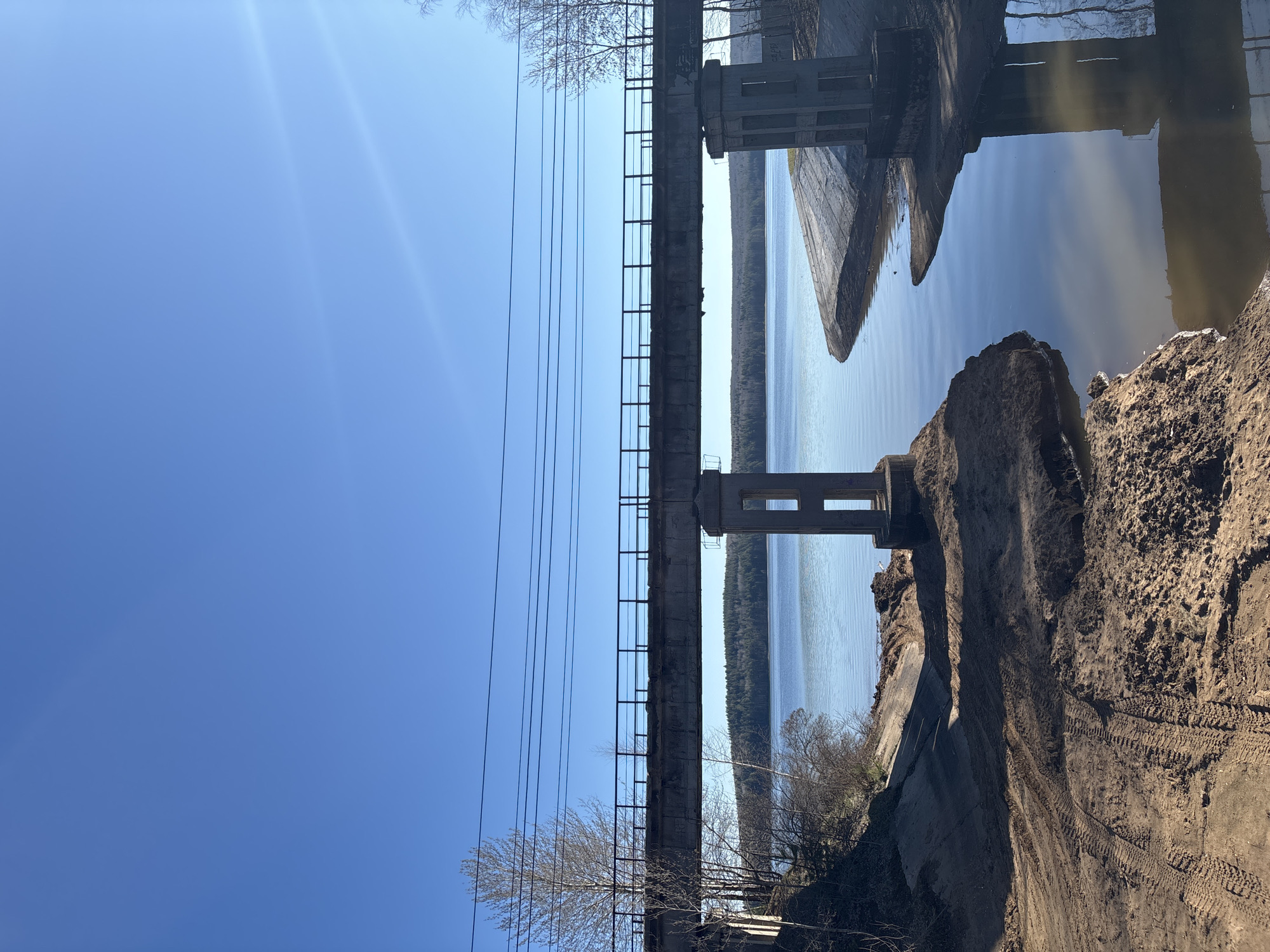 The image depicts a concrete bridge or overpass, likely a structure of some age given its weathering and design, over a body of water. The setting appears to be a natural environment, likely near the city of Chaikovsky, Russia, given the location tag. The foreground shows an eroded dirt bank with tire tracks, suggesting recent construction or earth-moving activities. The water is calm and reflects the clear blue sky, creating a mirror-like effect. Power lines run alongside the bridge. The overall scene is a mix of industrial and natural elements.