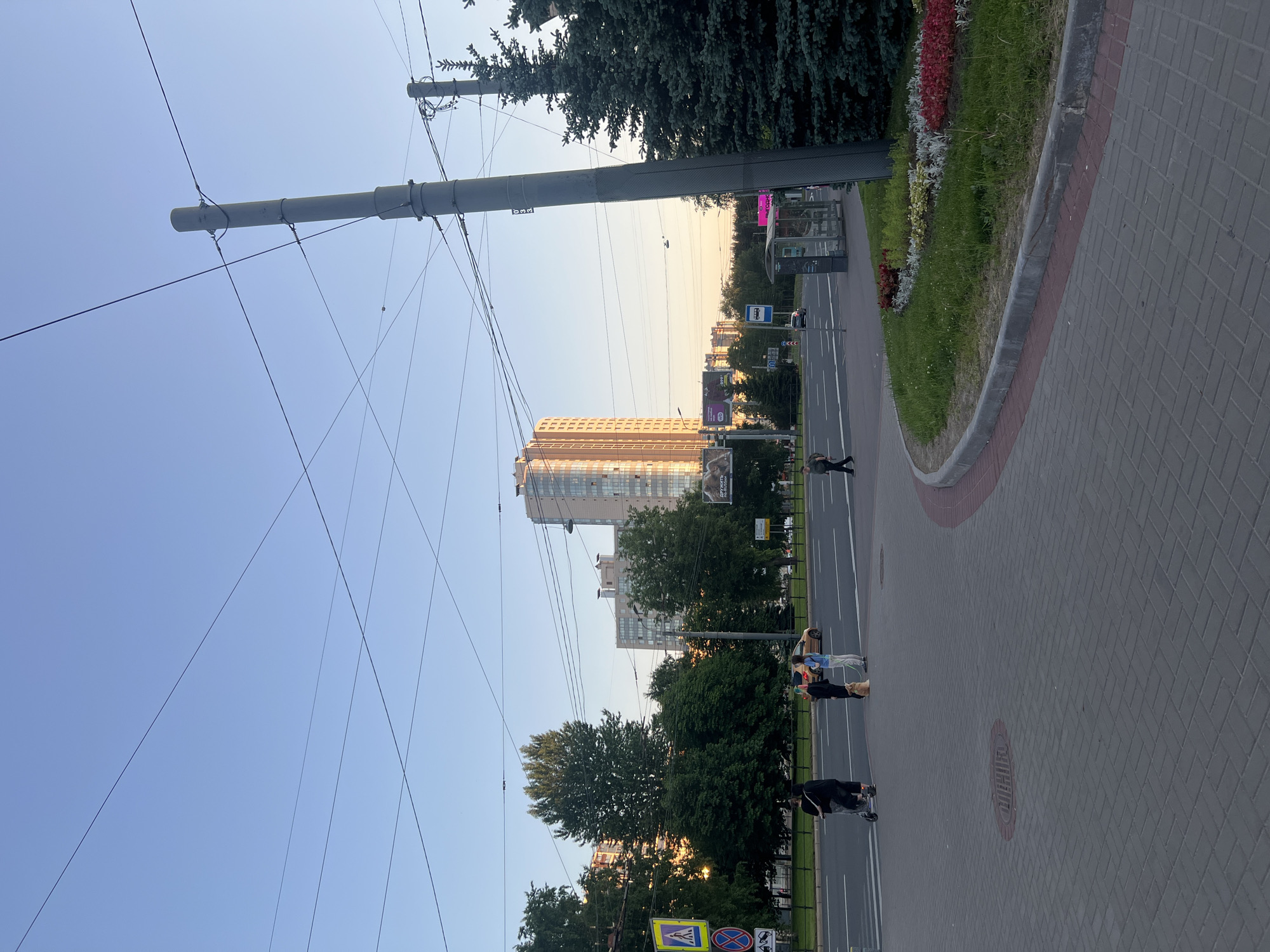 This image depicts an urban street scene in an unknown city in Russia during late afternoon or early evening, characterized by a clear sky and the warm light of a low sun.
The upper portion of the frame is dominated by a clear, pale blue sky, transitioning to a lighter yellowish-orange hue near the horizon. Numerous thin, dark overhead utility wires crisscross the sky.
In the foreground, a grey paver sidewalk, laid in a herringbone pattern, occupies the lower half of the image. A circular manhole cover is embedded in the pavement on the left side of the sidewalk. To the right, a curved landscaped area with grass and patches of red, white, and yellow flowers is bordered by a concrete curb.
On the right side, a tall, grey concrete utility pole, supporting multiple black cables and wires, stands prominently. Behind it are several dark green coniferous trees. A dark grey bus stop shelter with translucent panels is positioned on the sidewalk to the right.
On the left side of the frame, a wide, multi-lane asphalt road with white markings runs horizontally. Four individuals are present on or near the sidewalk adjacent to this road. From left to right: one person in dark clothing is operating an electric scooter; two individuals stand side-by-side, one in dark attire and the other in a light blue top and light pants, holding a leash attached to a light brown dog positioned between them. Further across the street, a lone figure in dark clothing walks away from the camera.
Road signage on the left includes a yellow square pedestrian crossing sign and a red circular "no stopping" sign. In the middle ground, beyond the road and a line of green deciduous trees, multiple tall buildings are visible. A prominent high-rise building with a light-colored facade reflects the sunset, appearing golden-orange. A large billboard with Cyrillic text is partially visible to its left. Other buildings and distant greenery are discernible under the horizon. Numerous overhead wires, consistent with a trolleybus or tram system, span across the street.