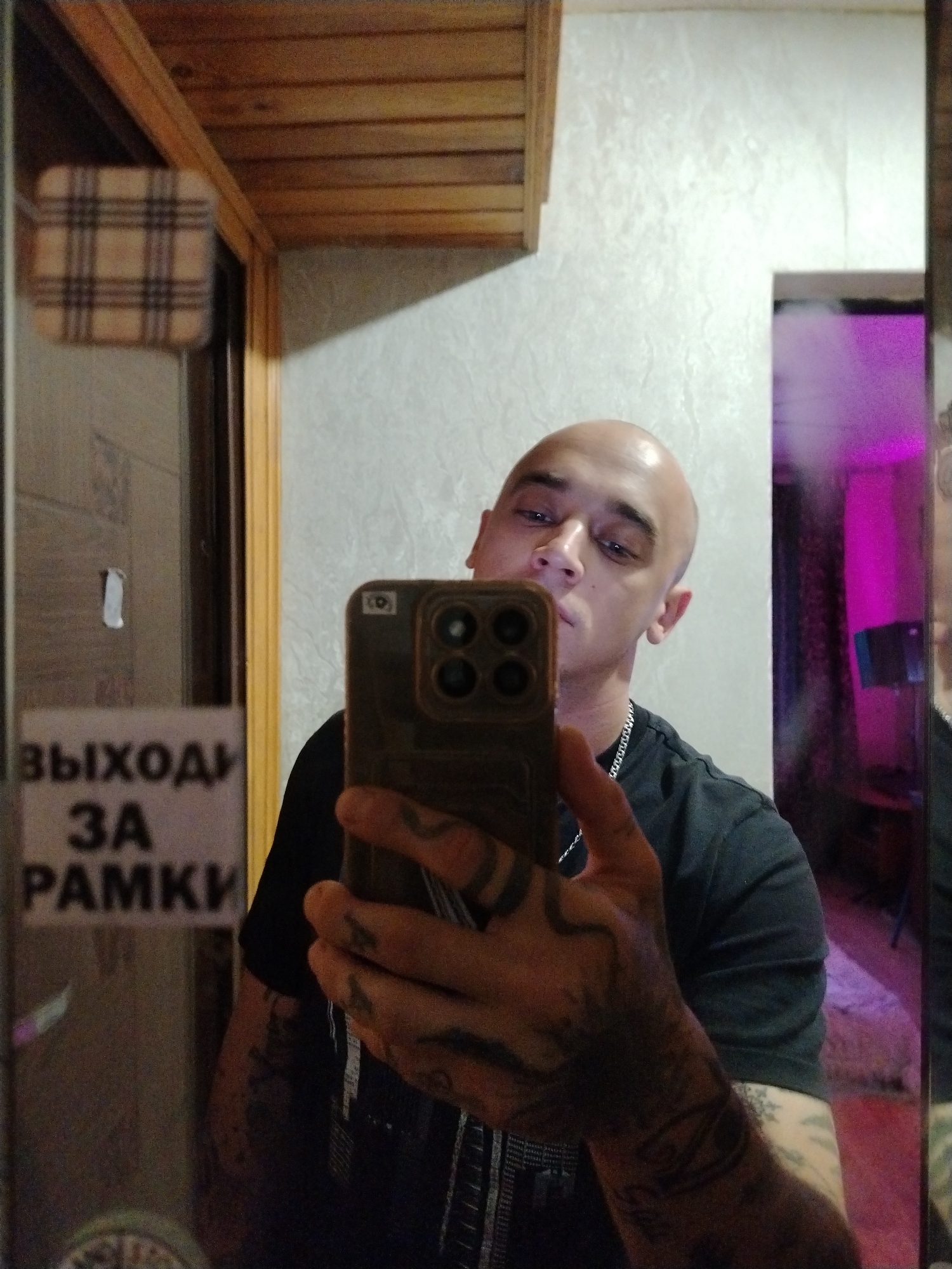 A bald man is taking a selfie in a mirror. He is wearing a black t-shirt and has tattoos on his arms and hands. The background shows a wall with a wooden ceiling and a sign with Russian text that appears to read "ВЫХОДЫ ЗА РАМКИ" which translates to "EXITS BEYOND THE FRAMES". There is also a reflection of a room with pink lighting.