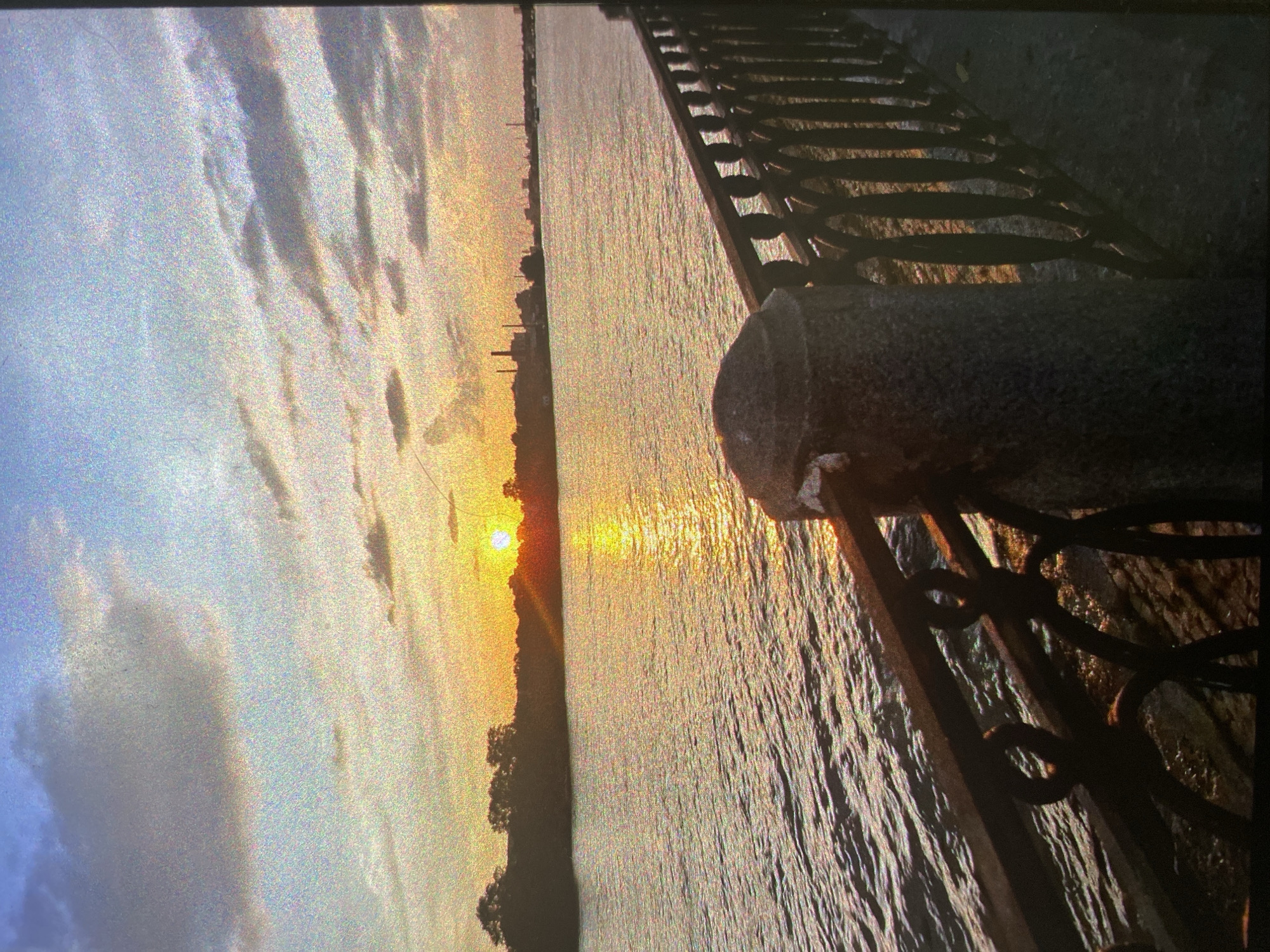 The image depicts a tranquil sunset scene over a wide body of water in Saint Petersburg, Russia. The view is taken from an embankment, with a decorative metal fence or railing running along the right side of the foreground. This fence features intricate, curvilinear patterns between its vertical supports, with a prominent stone or concrete pillar anchoring it in the bottom right corner.
The sun is positioned low on the horizon, casting a brilliant, golden light that reflects intensely across the surface of the water, creating a shimmering path. On the distant bank, the silhouette of a city skyline, interspersed with trees, is visible against the fading daylight. The sky transitions from soft blues and grays on the left to warm oranges and yellows near the setting sun, with scattered clouds illuminated by the golden light.
No people are visible in the frame. The scene captures the peaceful end of the day, with the calm water and colorful sky evoking a sense of serenity. The image possesses a slightly grainy texture, contributing to its atmospheric quality. There is no visible text.