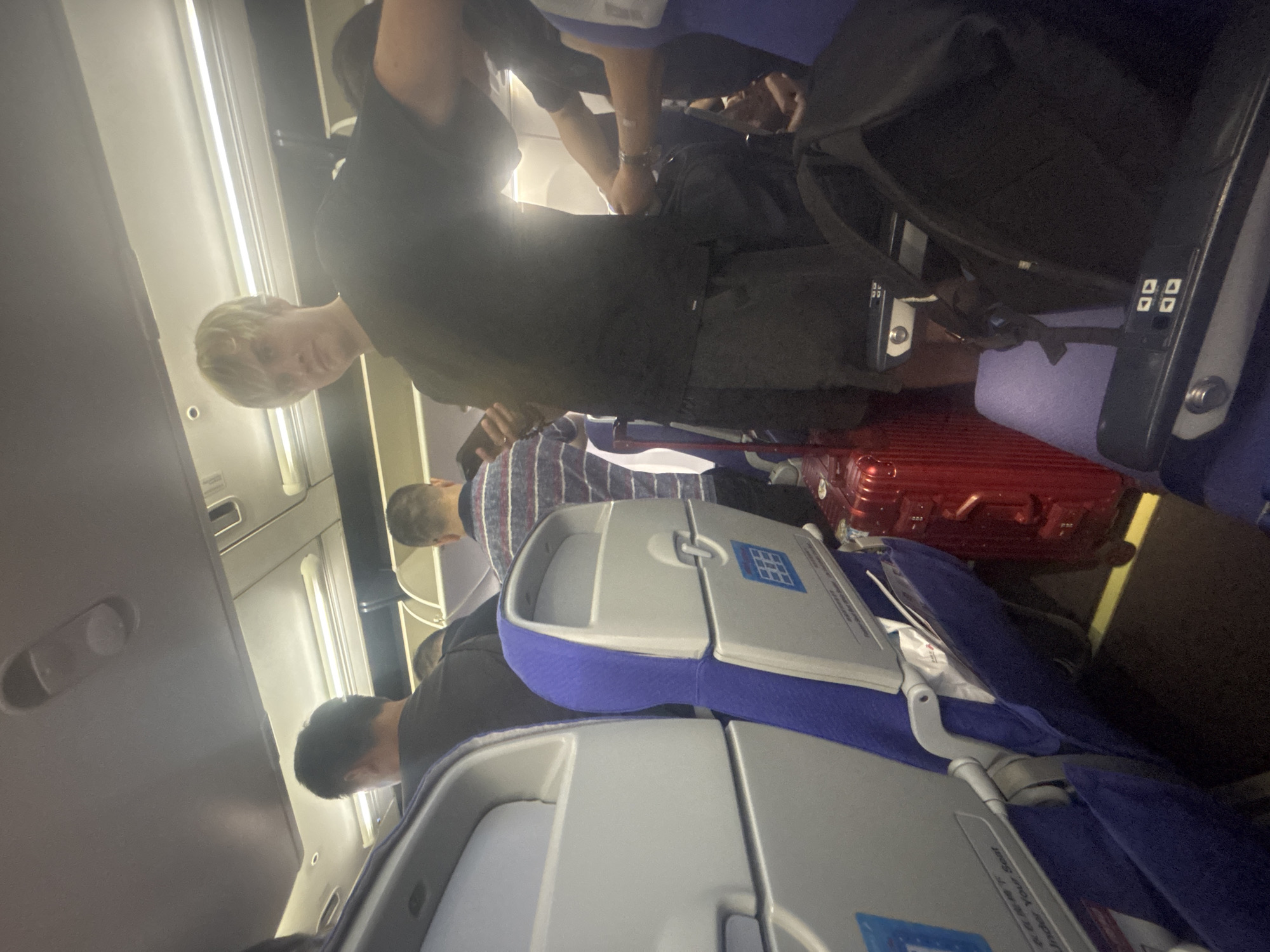 The image depicts the interior of an airplane cabin, likely in flight. The scene is crowded, with several passengers visible in the aisle and seated. In the foreground, a young man with blond hair, wearing a black t-shirt and dark shorts, is looking towards the camera and holding a smartphone. To his left, a red suitcase is placed in the aisle, blocking passage. Other passengers are seen further back, some appearing to be standing or moving within the confined space. The seats are upholstered in blue and gray fabric. The overhead compartments and lighting fixtures of the airplane are visible at the top of the frame. The specific location in Xinwu District, China, cannot be determined from the image alone.