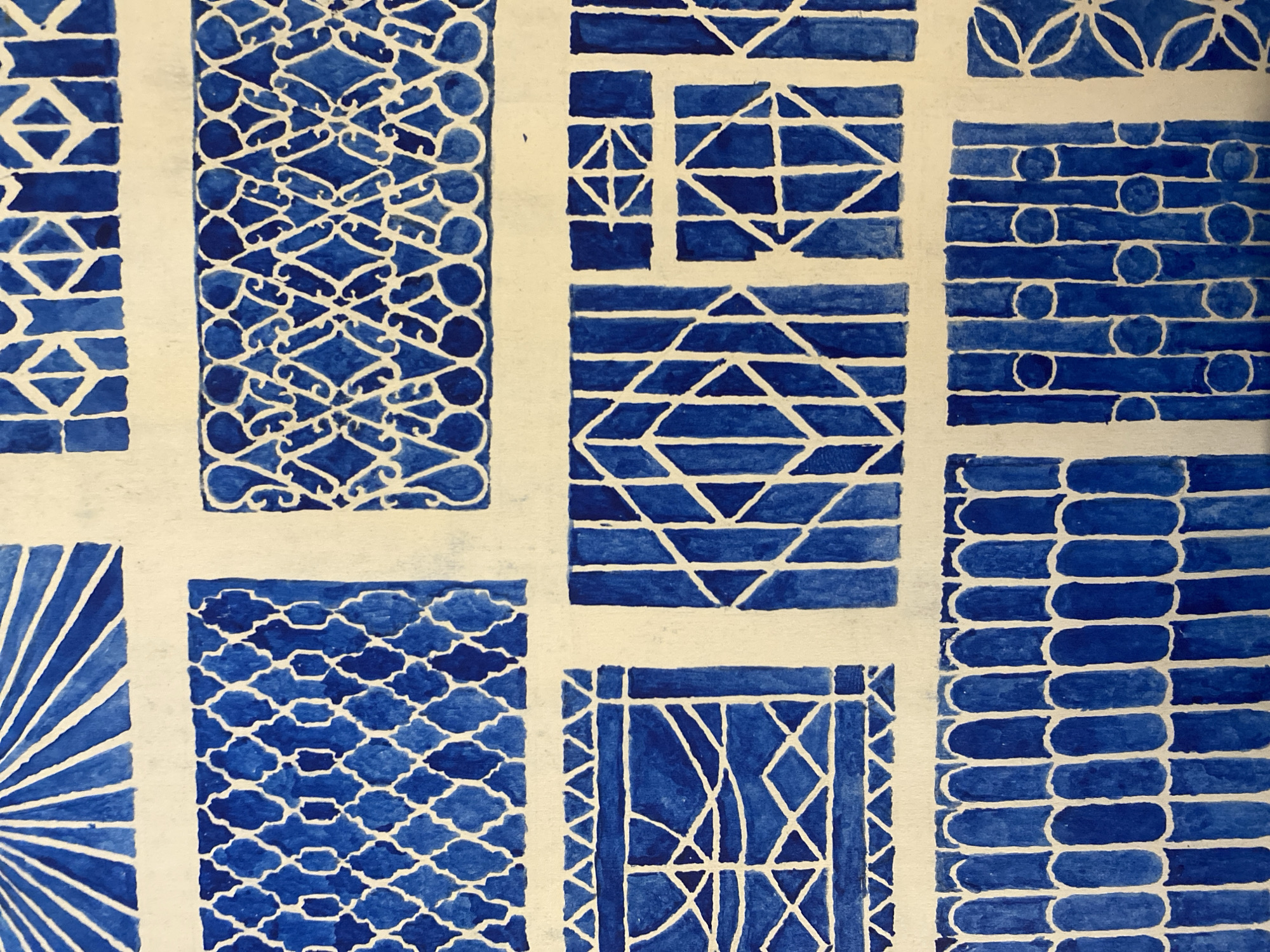 This is a close-up, overhead shot of a watercolor painting on textured paper. The painting is abstract, featuring a repeating pattern of blue and white geometric shapes and motifs. There are no people, objects, or discernible scenes depicted, only abstract patterns.
The painting is divided into several rectangular sections, each with a distinct design. Some sections contain rows of interlocking diamond shapes, others feature parallel lines forming angular patterns, and some have repeating circular or oval elements. The blue paint is applied in varying shades, with some areas appearing darker and more saturated than others, suggesting the use of water to create lighter tones. The white areas are the unpainted paper, highlighting the negative space within the patterns.
The style of the painting is reminiscent of traditional tile work or decorative motifs, with the blue and white color scheme evoking imagery of Delft pottery or Chinese porcelain. The texture of the paper is visible, adding to the tactile quality of the artwork. There is no text visible in the image. The lighting appears to be neutral, with no strong shadows, suggesting an indoor setting or diffused natural light. The overall impression is one of intricate, stylized design.