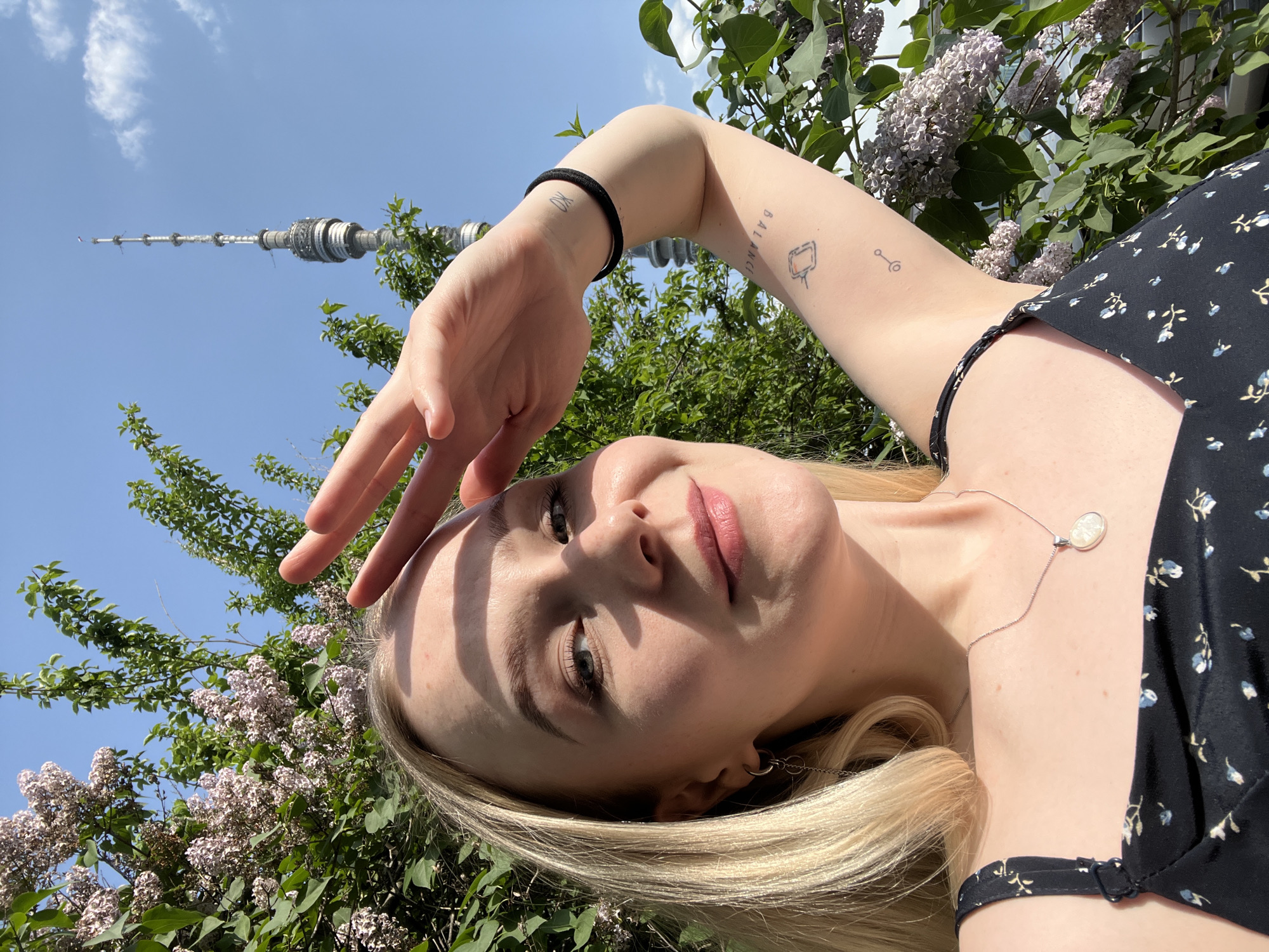A young woman is photographed from a low angle, looking up toward the camera. She is shielding her eyes from the sun with her left hand. She has fair skin, light blonde hair, and a necklace with a pendant. She wears a black floral-patterned top. Tattoos are visible on her left forearm. In the background, there is a blue sky, green foliage, and lilac flowers. The Ostankino Tower in Moscow, Russia, is visible in the distance. The scene appears to be set in a park in the city of Khimki, Russia.