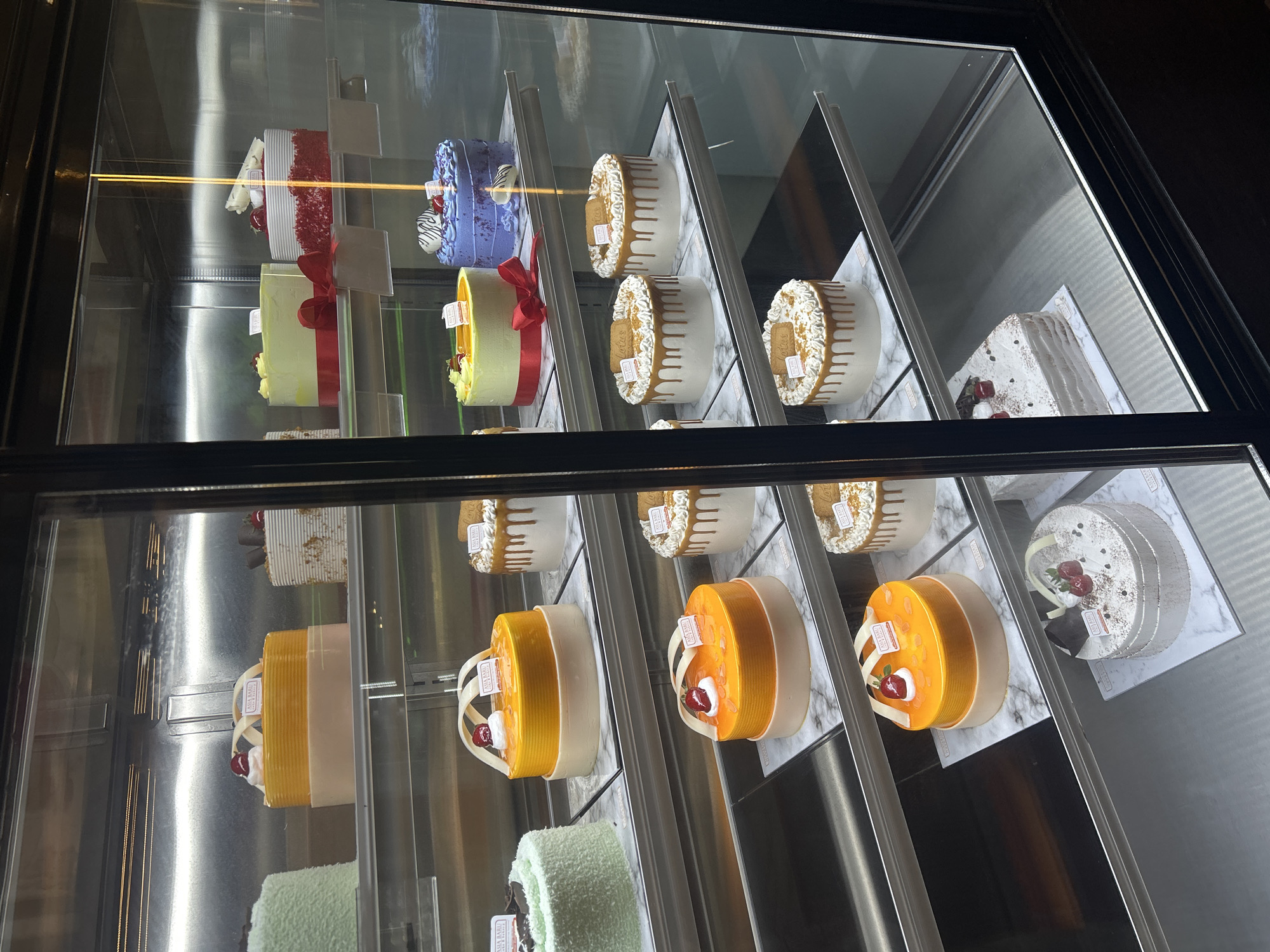 A display case in a bakery in Makassar, Indonesia, contains various cakes. The cakes are arranged on multiple shelves, each placed on a marble-patterned stand. The display case has glass doors, reflecting the interior lighting.
On the top shelf, from left to right, are a light green cake, a yellow cake with a beige base and a cherry on top, a white cake with brown speckles and a cherry, a light green cake with a red ribbon, and a red velvet cake with white frosting and white chocolate decorations.
The second shelf from the top features a green cake with dark speckles, an orange cake with a beige base and a cherry on top, a blue cake with white frosting swirls and cherries, and a white cake with dripping caramel and lotus biscoff cookies. Another similar white cake with caramel drip and cookies is next to it.
The third shelf has two orange cakes with beige bases and cherries on top. To their right, there are two white cakes with dripping caramel and lotus biscoff cookies.
The bottom shelf contains a white cake with layered frosting and chocolate decorations, and a white cake with square layers and frosting. There are also empty shelves and reflections visible. The overall impression is of a well-stocked bakery display.