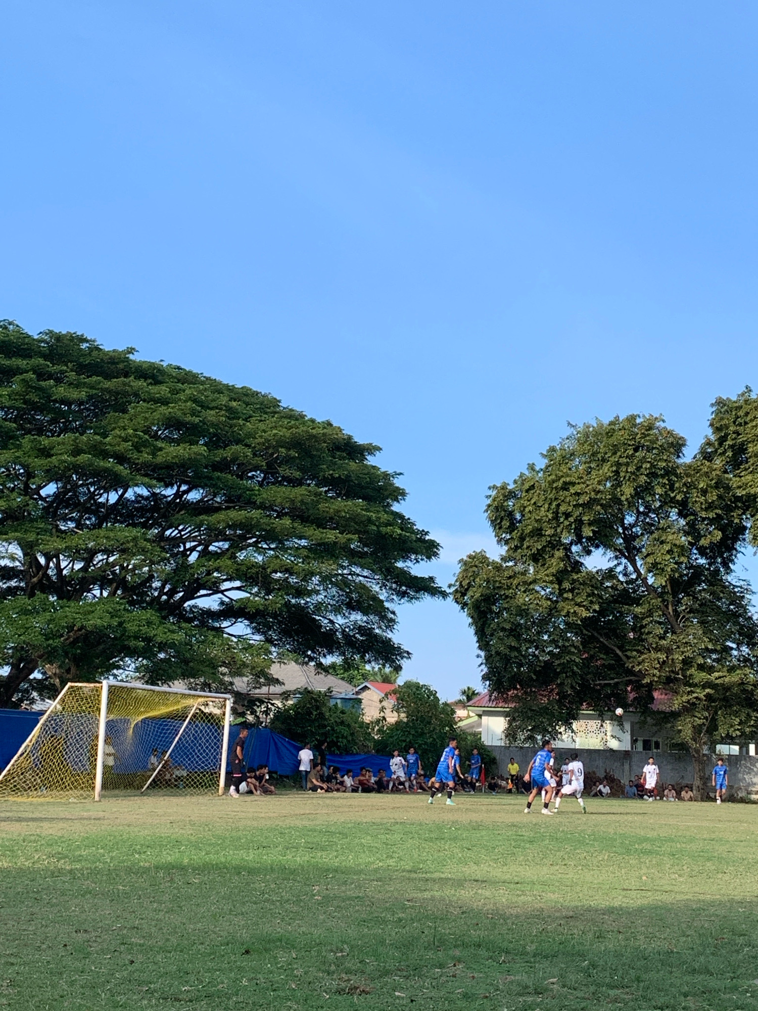 The image depicts a soccer game on a grass field in Keudelapang, Indonesia. A bright blue sky dominates the upper portion of the frame. To the left, a large, mature tree with dense green foliage is prominent, with a yellow and white soccer goal visible in the foreground. In the center and right of the field, several players in blue and white uniforms are actively engaged in the game, with one player in a white uniform with the number '9' visible. Spectators are seated and standing on the sidelines, many under a blue tarp for shade. Several houses with red roofs are visible in the background, partially obscured by trees. The overall environment suggests a warm, sunny day for an outdoor sporting event.