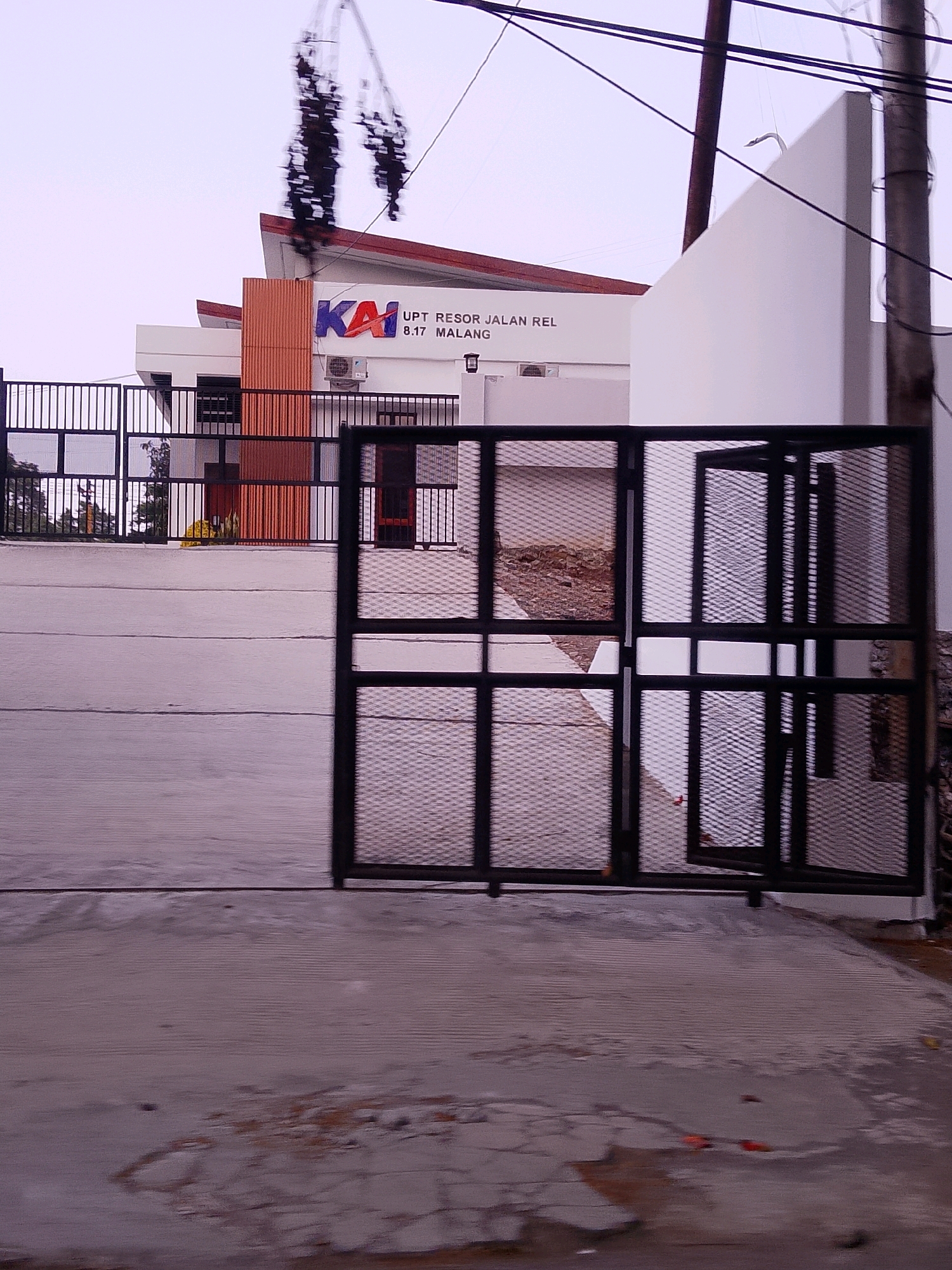 The image shows the facade of the UPT RESOR JALAN REL B.17 MALANG, a building in Malang, Indonesia. The building has a modern architectural style with white walls and an orange vertical accent panel. The KAI logo, which stands for Kereta Api Indonesia (Indonesian Railway Company), is prominently displayed on the building. In front of the building, there is a black metal gate with diamond-shaped mesh. Beyond the gate, a concrete pathway leads to the building entrance. The sky is overcast, and there are some power lines visible in the upper part of the image. The overall scene suggests an administrative or operational building related to the railway system.
