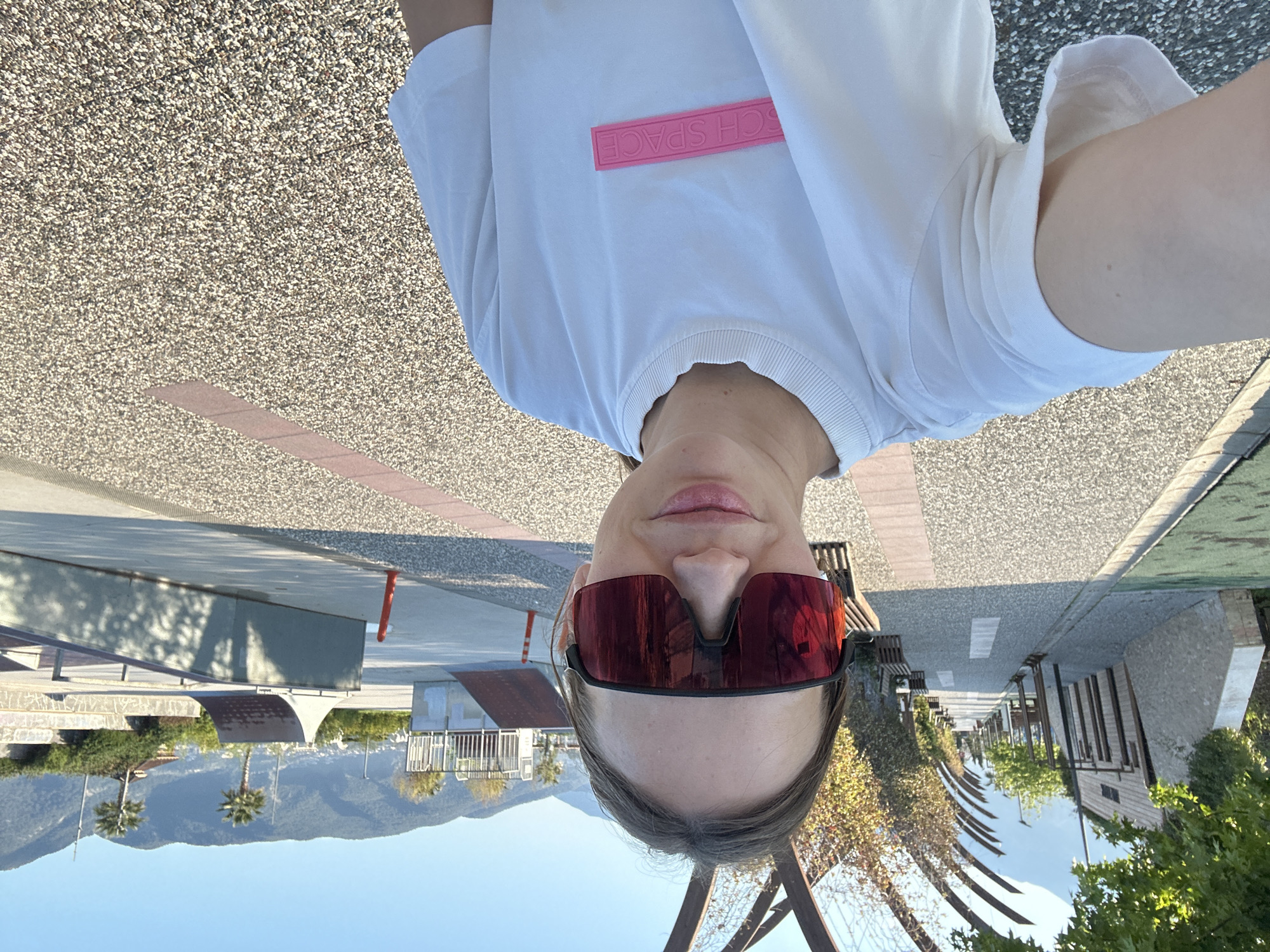 A person is taking a selfie in Fethiye, Turkey. The individual wears red sunglasses and a white t-shirt with a pink rectangular design on the chest. The background includes a skate park, mountains, palm trees, benches, a sidewalk, and other street furniture. The sky is blue. The perspective is upside down.