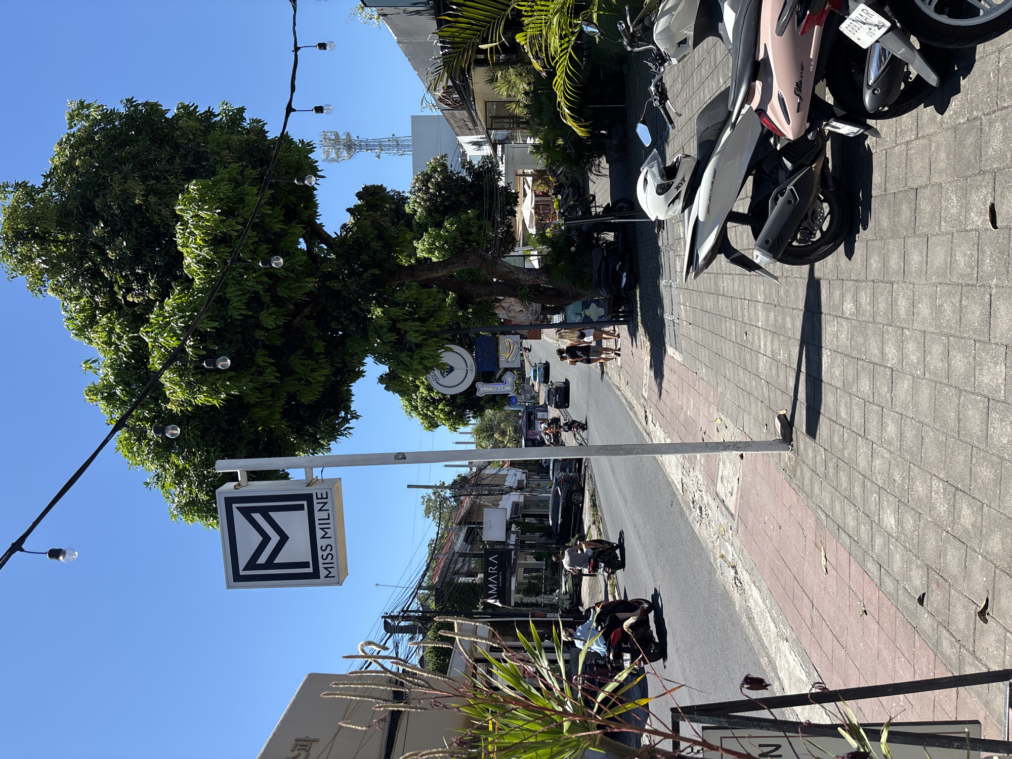 The image shows a street scene in Pecatu, Indonesia. The photo is taken from a slight elevation, looking down a street with a mix of pedestrians, parked motorbikes, and passing traffic. A sign for "MISS MILNE" is prominently displayed on the left side of the frame. Lush tropical foliage is visible, including a large tree and various plants along the street. The sky is clear and blue. Power lines and a cell tower can be seen in the background. On the right, several motorbikes are parked on the sidewalk. A store sign for "AMARA" is visible on the buildings. Several people are walking on the sidewalk.