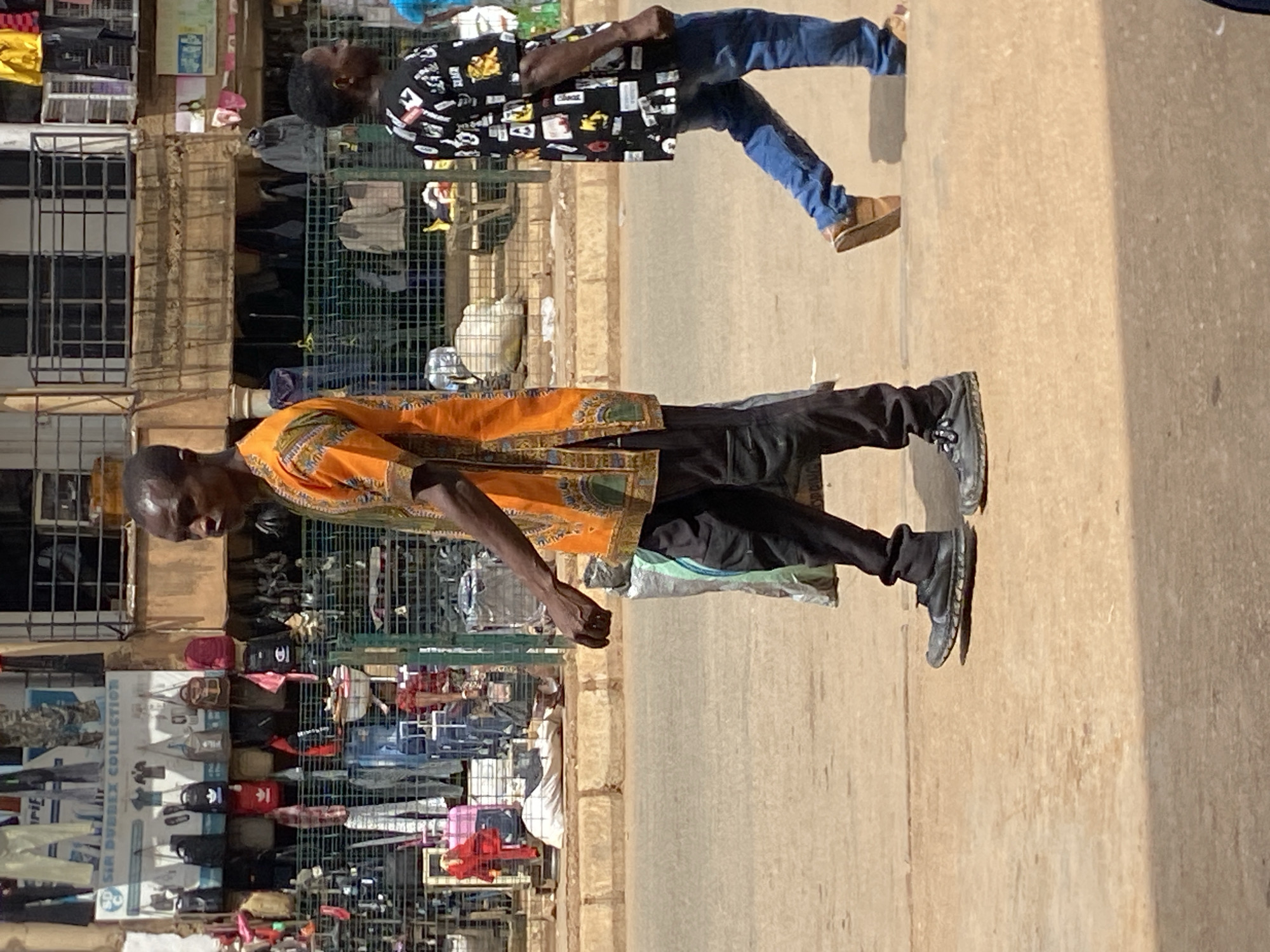An outdoor scene in Bukuru, Nigeria, shows two men on a paved or dusty street, likely within a market area, under bright daylight. The image is oriented vertically, showing the subjects and surroundings rotated 90 degrees clockwise.
In the foreground (when rotated correctly), a man with dark skin and short dark hair stands looking towards the left. He is wearing a vibrant orange and green patterned long-sleeved shirt with black pants and black sneakers. His left arm is slightly bent, and his right arm is partially visible.
Further back, towards the right, another man with dark skin and short dark hair is visible. He is wearing a black long-sleeved shirt adorned with various white and yellow patterns and graphics, blue jeans, and light brown shoes. He also appears to be standing or casually walking, looking in a similar direction as the foreground man.
Behind the men, the background reveals a bustling market setting. There are various stalls and shops displaying merchandise, including clothes hanging on racks and shelves, and bags. One prominent display board features the text "SIA DUBEX COLLECTION" and logos resembling those of sports brands like Adidas and Nike. Buildings with metal-grated windows are visible behind the market stalls. The overall scene suggests a vibrant commercial street or market in sunny weather, with strong shadows indicating the mid-day sun.