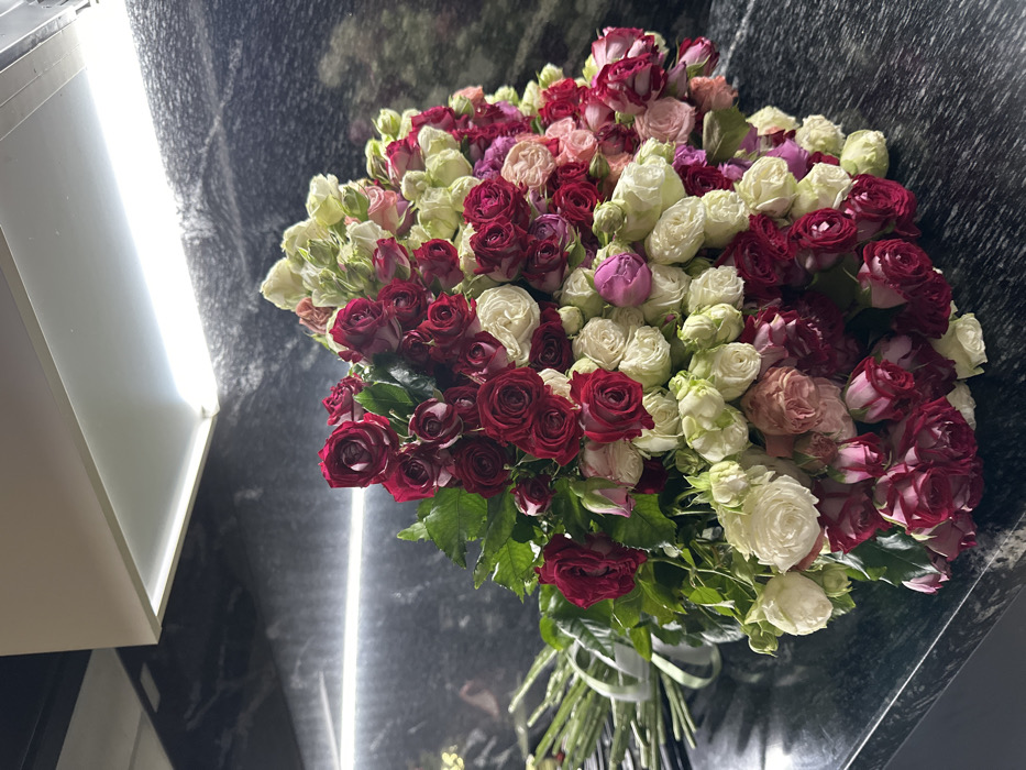 A lush bouquet of roses, predominantly deep red and creamy white, with hints of pink and purple, occupies the frame. The roses are tightly packed, forming a dense, voluminous arrangement. Green stems and leaves are visible near the bottom, bound by a light-colored ribbon. The background is a dark, mottled surface, possibly marble, with bright, linear light reflections. A white, angular structure is present on the left side of the image.