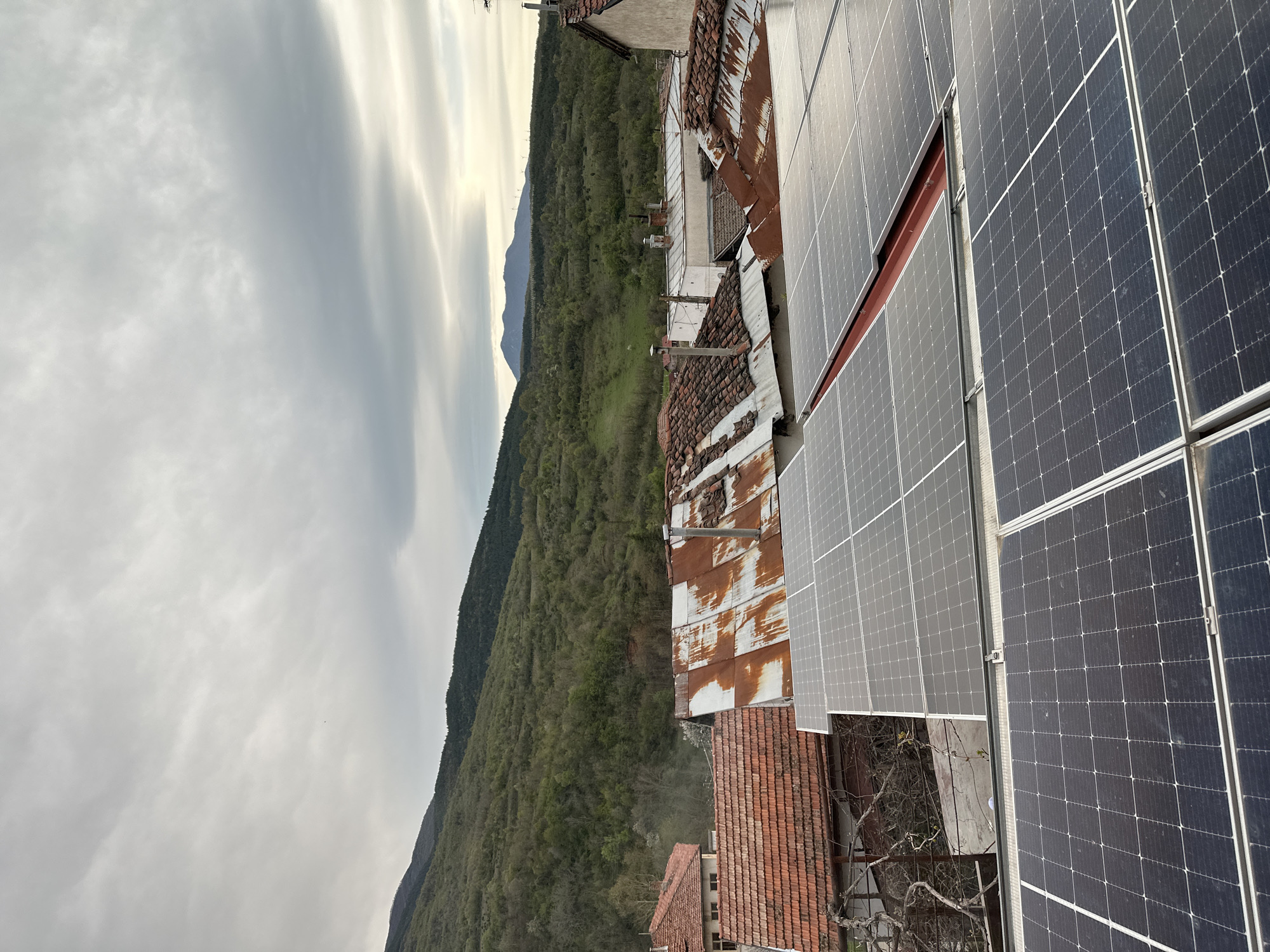 The image shows a view from a rooftop in what appears to be Petrovo, Bulgaria, judging by the architecture and mountainous terrain. The foreground is dominated by rows of solar panels, with some appearing cleaner and newer than others. Adjacent to the solar panels are older buildings with rusted metal roofs and tiled roofs of varying conditions. Beyond the buildings, a heavily forested mountain range stretches into the distance under an overcast sky. The overall scene suggests a juxtaposition of older and newer technologies in a rural or semi-rural setting.