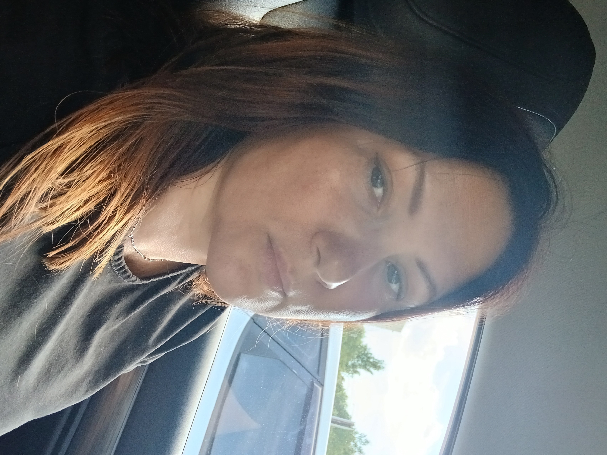 The image is a close-up, sideways portrait of a woman's face. She has dark hair with lighter, reddish-brown highlights, and her hair is styled loosely around her face. Her skin appears to be fair with some slight redness, and she is wearing a dark gray t-shirt with a ribbed neckline. A delicate silver necklace is visible around her neck. Her expression is neutral, and her gaze is directed slightly away from the camera.
The background suggests she is in a car, indicated by the car window and the interior of the vehicle. Through the window, trees and a bright sky with some clouds are visible, hinting at an outdoor setting during the daytime. The lighting is somewhat harsh, suggesting it might be a sunny day. The overall mood is casual and unposed. No visible text is present in the image.