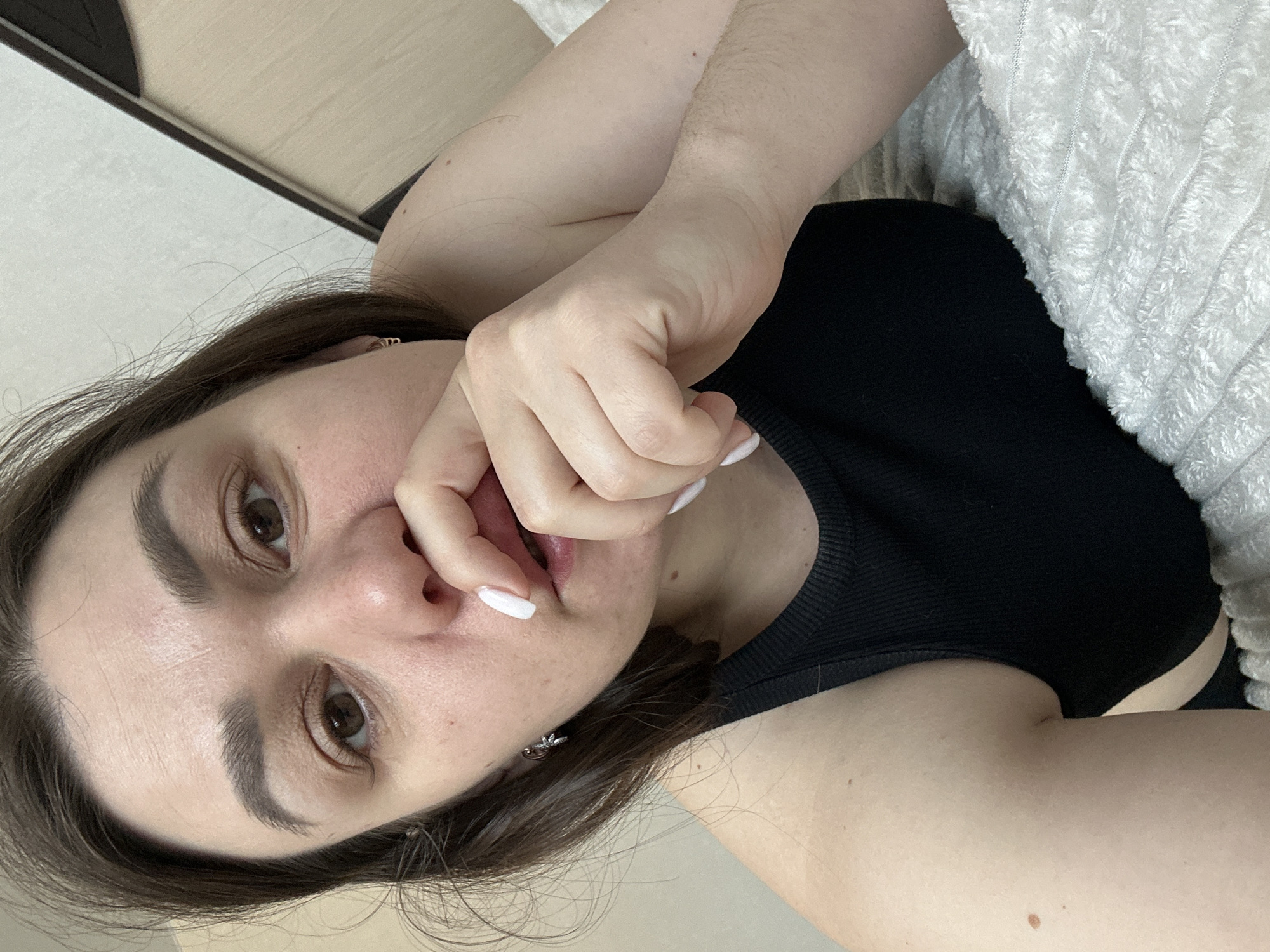 A woman with brown hair and eyes is lying down, wearing a black top. Her right hand is near her mouth. The background includes a beige wall and a white blanket. The location is stated to be in Chaikovsky, Russia, but there are no elements visible in the image that confirm that information.