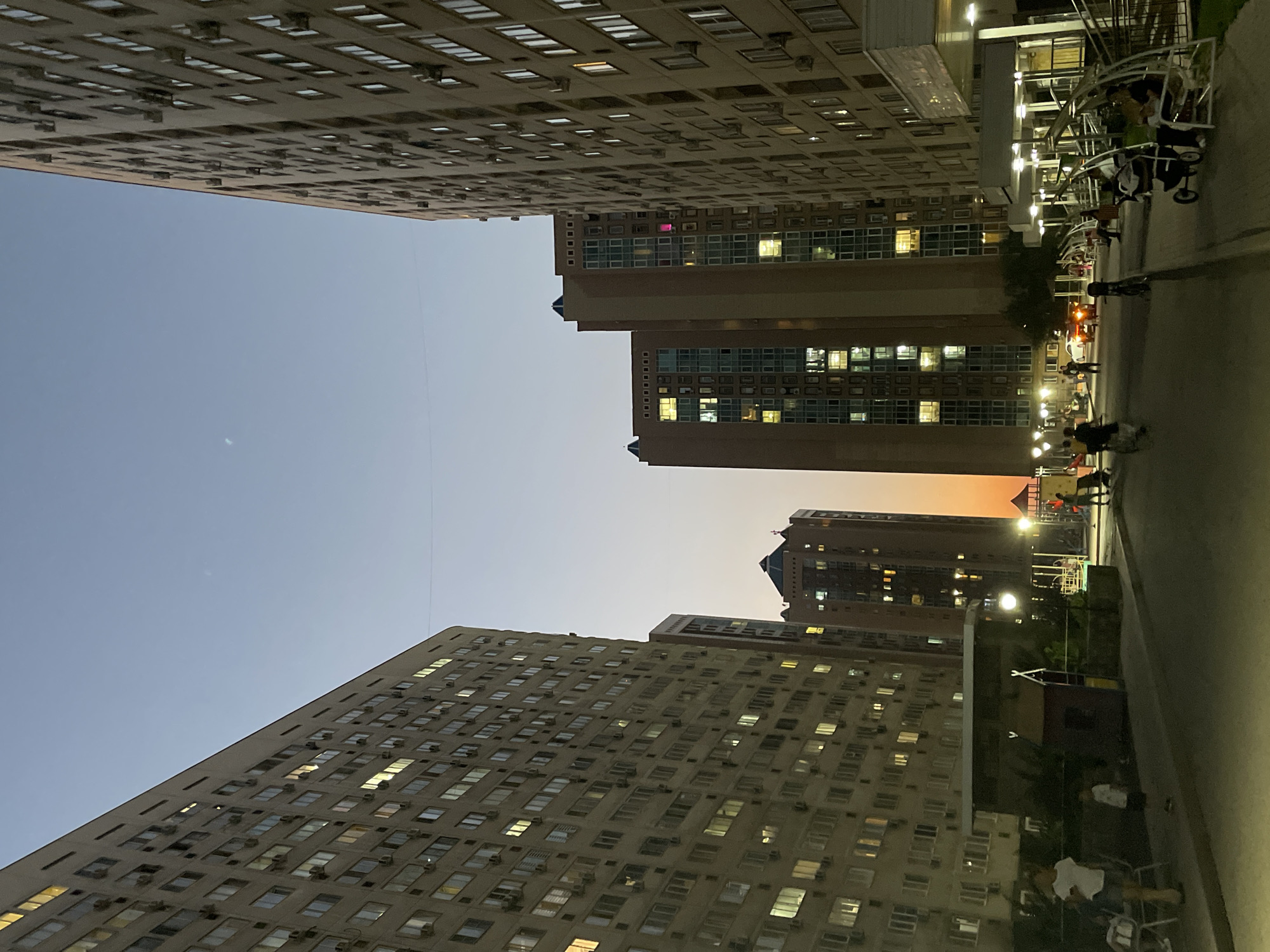 The image shows an evening scene in a residential complex in Almaty, Kazakhstan. Two large, light-colored apartment buildings dominate the left and right sides of the frame, with numerous windows illuminated from within. In the background, two taller, darker buildings rise, also showing illuminated windows. The sky above is a gradient of pale blue and orange, indicative of twilight.
In the foreground, a paved walkway and street are visible. Several people are present: a woman and child are sitting on chairs to the left, a child stands near a small play structure in the middle ground, and a group of people are walking and sitting on benches in the right foreground, some with strollers. A few cars are parked or driving in the background. The overall atmosphere is one of a typical urban residential area at dusk.