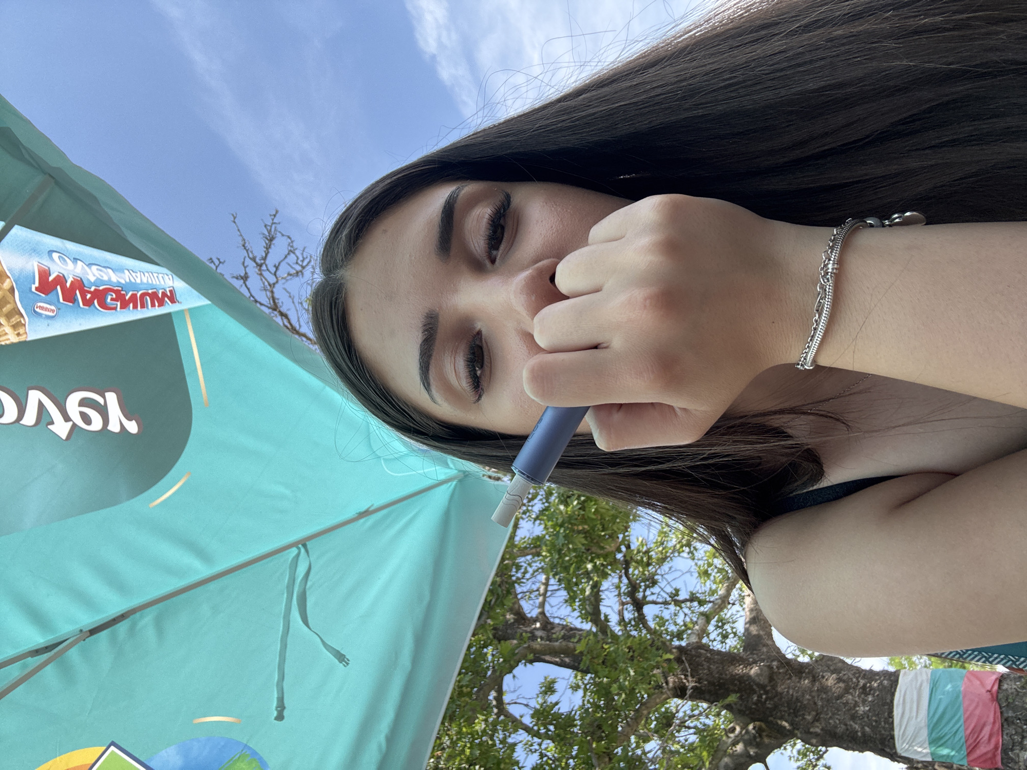 An outdoor portrait shot in Kostievo, Bulgaria, features a young woman positioned centrally under a large teal umbrella or canopy. The woman has long, dark-brown hair and dark eyes, looking directly at the camera. Her left hand is raised, partially obscuring her nose and mouth, and she holds a blue cylindrical object, consistent with a vape pen, with a white tip near her face. She wears a dark-colored top and a silver multi-strand bracelet with charms on her left wrist. The teal umbrella, which covers the upper portion of the frame, displays inverted and mirrored branding, including "MAGNUM" clearly visible on an ice cream cone graphic, and other indecipherable text. In the background, under a clear blue sky with sparse white clouds, are large trees with dense green foliage and thick trunks. One tree trunk in the lower-left is adorned with horizontally wrapped fabric strips in white, green, and red.