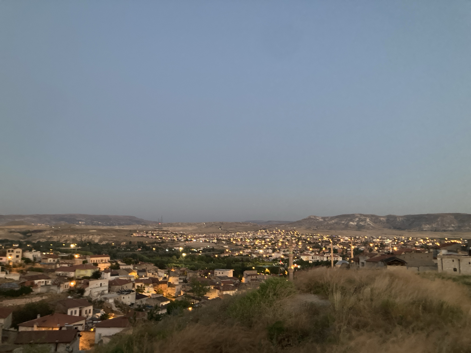 This is a landscape photograph taken at dusk, overlooking the city of Avanos İlçe Merkezi, Turkey. The foreground is dominated by dry, tall grass and some rough terrain. In the mid-ground, a dense collection of buildings with red-tiled roofs is visible, with many windows and some structures illuminated by warm light. Further into the background, the city stretches out, characterized by a multitude of lights from houses and streetlamps, creating a warm glow. The horizon is marked by rolling hills and mountains, some with distinct geological formations. The sky above is a clear, pale blue, transitioning to a darker shade near the horizon, indicating the twilight hour. No people are visible in the frame. The overall scene depicts a quiet, illuminated urban settlement nestled within a natural, hilly landscape.