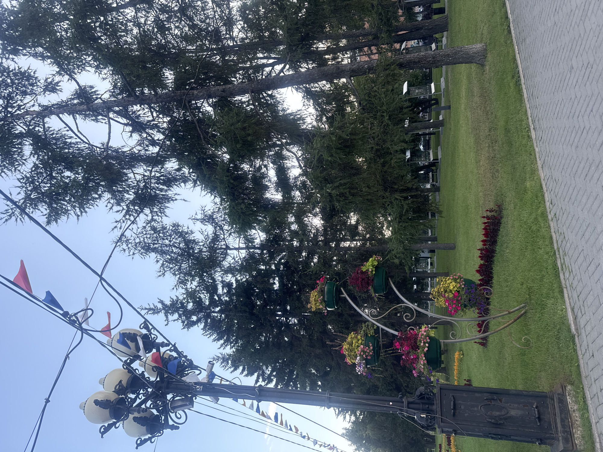 In Irkutsk, Russia, this image captures a park scene with a focus on a decorative lamppost and flower arrangements. The lamppost, made of dark metal, features multiple globe-shaped lamps and is adorned with small, colorful flags that appear to be red, white, and blue. A structure with several tiers of hanging planters, filled with a variety of colorful flowers, is positioned next to the lamppost. The background consists of a dense line of tall evergreen trees, likely larch or pine, with a clear sky overhead. The ground is covered in manicured green grass, and a paved walkway made of grey interlocking bricks is visible in the foreground. The overall impression is of a well-maintained public space, possibly decorated for a holiday or event.