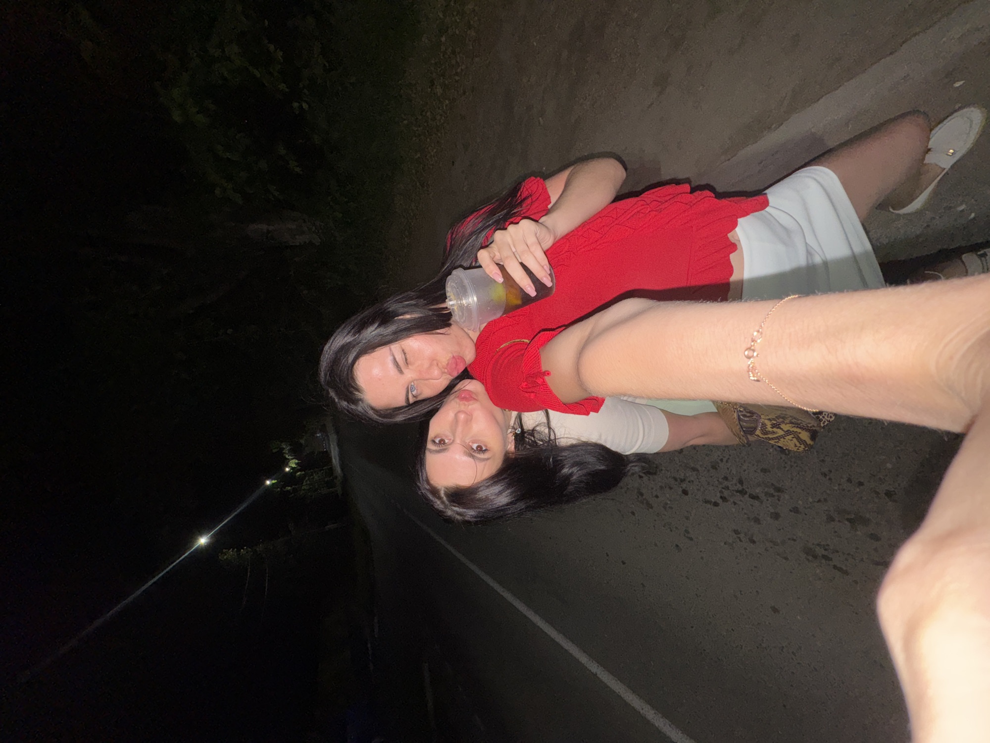 Two young women are posing for a selfie on a dark, paved road at night, possibly in Chaikovsky, Russia. The woman on the left, with dark hair, leans her head towards the woman on the right. The woman on the right, also with dark hair, is wearing a red top and a white skirt and is holding a drink in a plastic cup. She is winking at the camera. The road appears to be illuminated by distant streetlights.