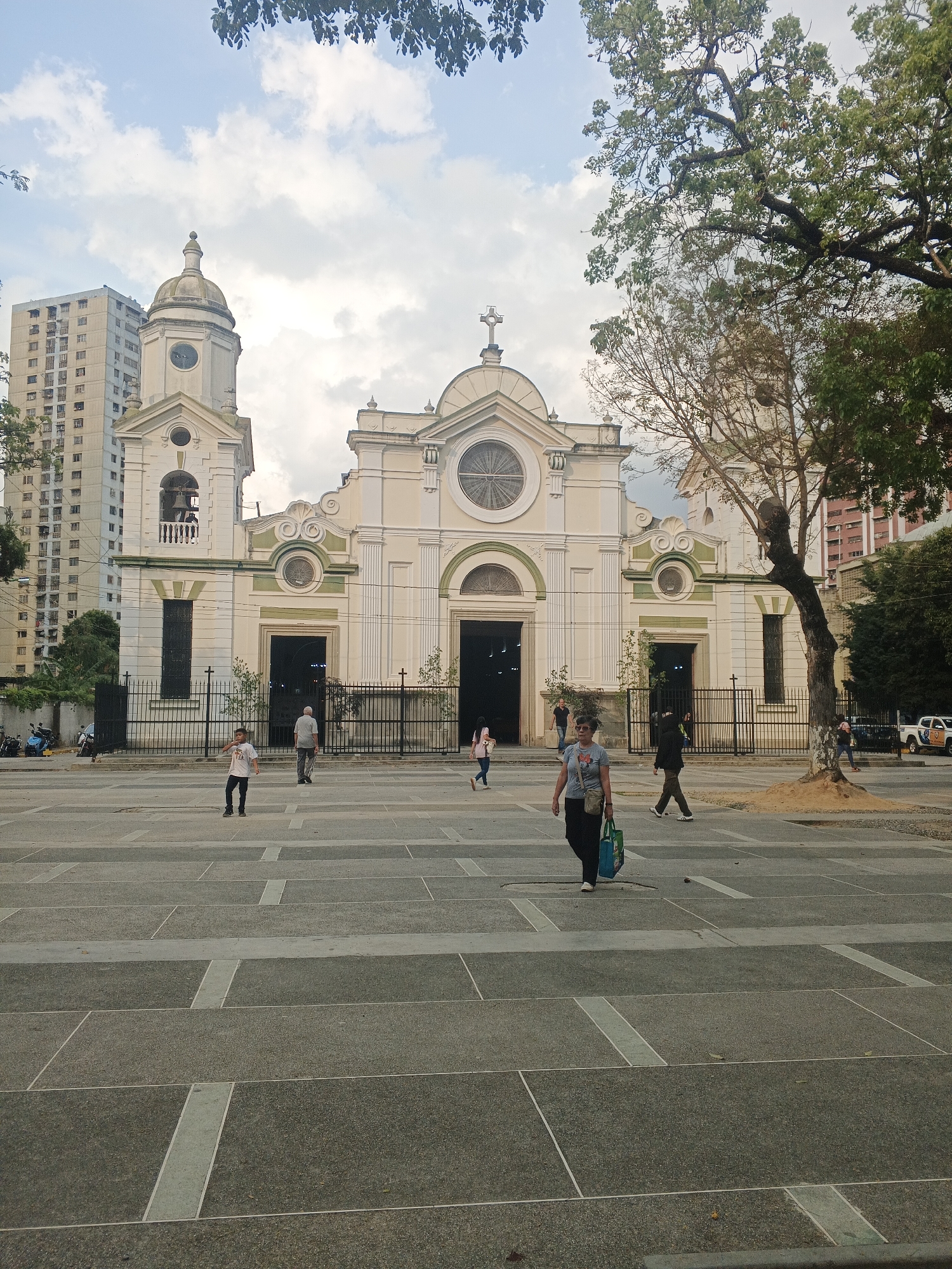 The image captures the Metropolitan Cathedral of Caracas in Venezuela, situated in Plaza Bolívar. The cathedral, a cream-colored building with green accents, features a prominent central circular window and a cross atop its facade. To the left stands a bell tower topped with a dome-shaped cupola.
In front of the cathedral, the plaza's patterned stone paving serves as a large open area with people walking across. A woman, carrying a blue bag, is walking in the foreground, while a young boy stands to the left of her. A man walks towards the cathedral. People are also visible in the background in proximity to the cathedral's entrances.
Buildings line the sides of the cathedral, with an apartment building on the left and a taller building on the right. Trees stand beside the buildings, one partially obscuring the right side of the cathedral. The sky is overcast with clouds.