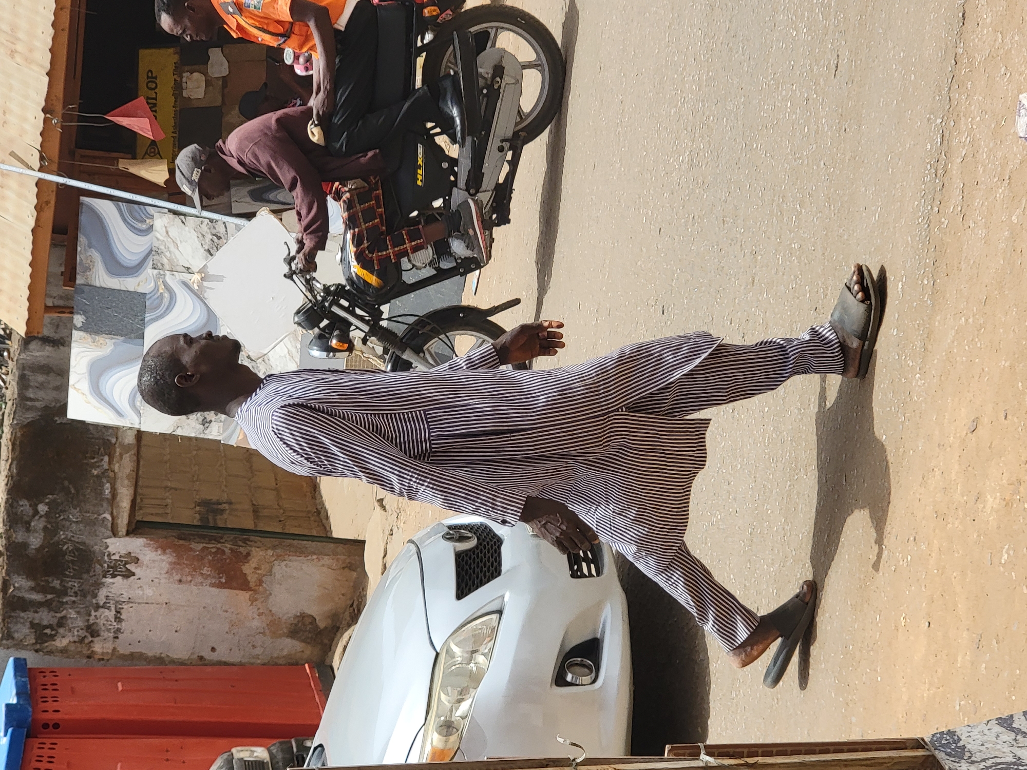 A man in a striped robe and sandals walks past a parked white car in Maiburiji, Nigeria. Two other men stand near a moped in front of a building. One is wearing an orange vest. The ground is sandy and the sky is clear.