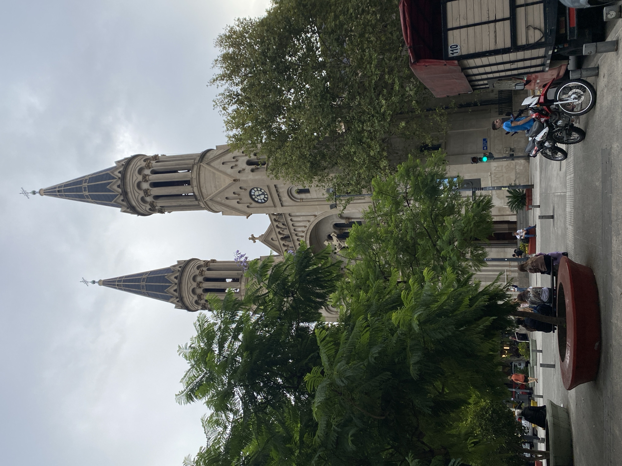 The image displays the exterior of the Parroquia Inmaculada Concepción, known as "La Redonda de Belgrano," located in Plaza Manuel Belgrano, within the Belgrano neighborhood of Buenos Aires, Argentina.
The central subject is the Neo-Romanesque church, constructed from light-colored stone, featuring two prominent octagonal bell towers topped with dark, pointed spires, each surmounted by a cross. The tower on the right (viewer's perspective) includes a large circular clock face indicating approximately 12:05. The church facade exhibits arched windows and decorative elements.
In the foreground, a public plaza with a paved surface is visible. It contains numerous trees with dense green foliage, including some exhibiting subtle purple blossoms, indicative of Jacaranda trees. Several concrete planters, some curved and grey, others circular with red rims, are scattered across the plaza.
Multiple individuals populate the scene:
* On the left, a person in dark attire is seated on a curved grey planter.
* Toward the center-right, a group of four people is seated on a bench behind a large red-rimmed planter.
* On the right side of the frame, a young adult male in a blue t-shirt stands beside two motorcycles: one red and white, the other black and white. He is positioned near a parked brown flatbed truck with wooden sides and a red tarp covering its load, displaying the number "110."
* Other pedestrians are visible walking or standing in the midground and background.
A green traffic light is mounted on a pole to the right of the church's entrance. The sky is uniformly overcast with grey clouds, suggesting daylight conditions with diffused illumination.