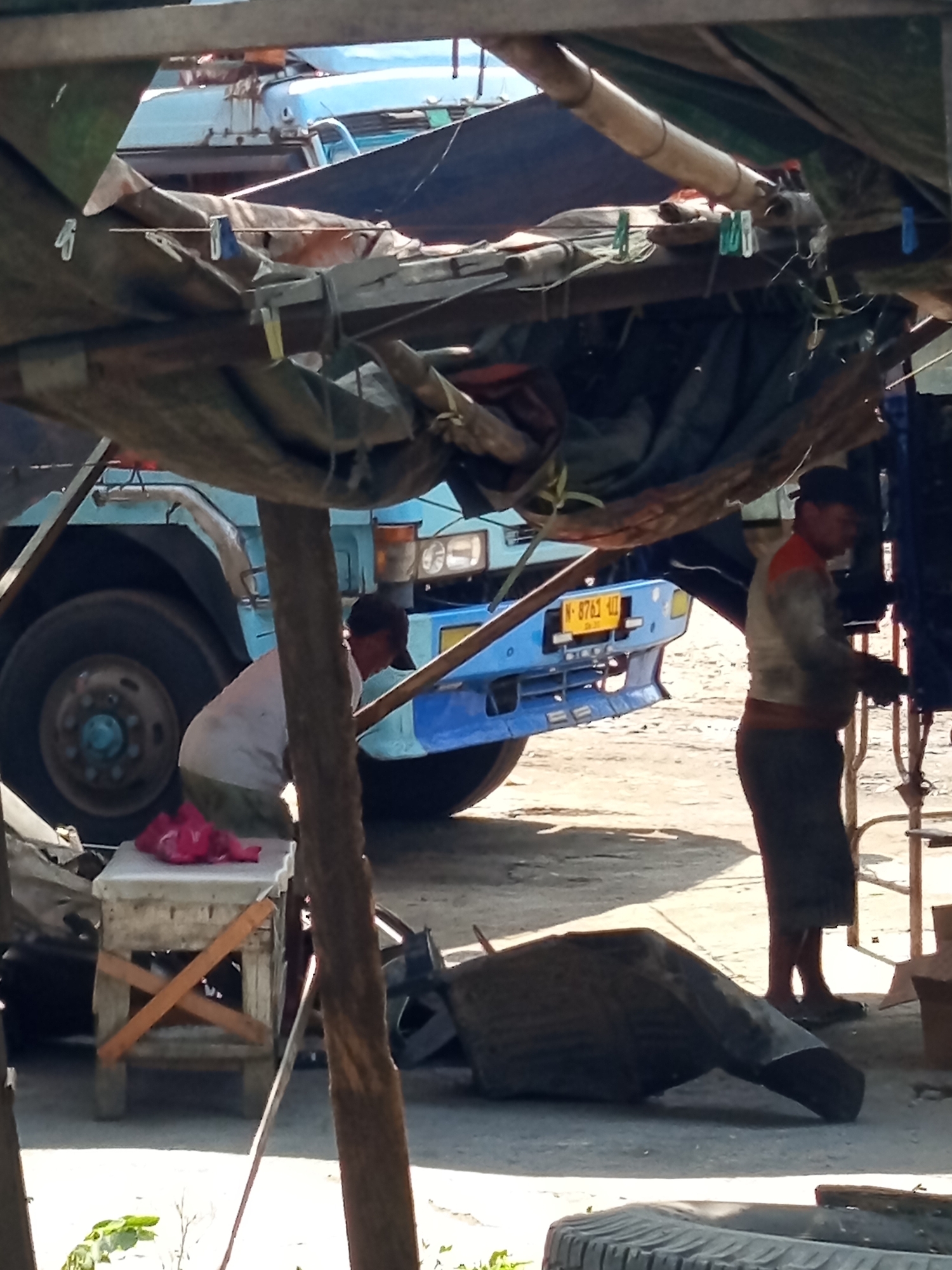 This is a medium shot, taken outdoors during the day, of a roadside repair or service area in Sidomulyo, Indonesia. The scene is cluttered with a variety of objects, suggesting a working environment. In the foreground, a small wooden stool with crisscrossed supports holds a bright pink plastic bag. To the right of the stool, a large, dark, tarp-covered object lies on the ground, possibly a piece of equipment or debris. Further back, a blue truck with a yellow license plate (N-8761-UJ) is parked. A man wearing a red and grey shirt and dark pants is standing next to the truck, appearing to be engaged in some activity, possibly repair work. Another person, wearing a white shirt and dark pants, is partially visible near the stool, also appearing to be working. The background shows more tarps and structures, possibly part of a makeshift shelter or workshop. The ground is dirt or gravel, indicative of an outdoor, possibly unpaved, location. The overall impression is one of an active, informal, and somewhat disorganized workspace.