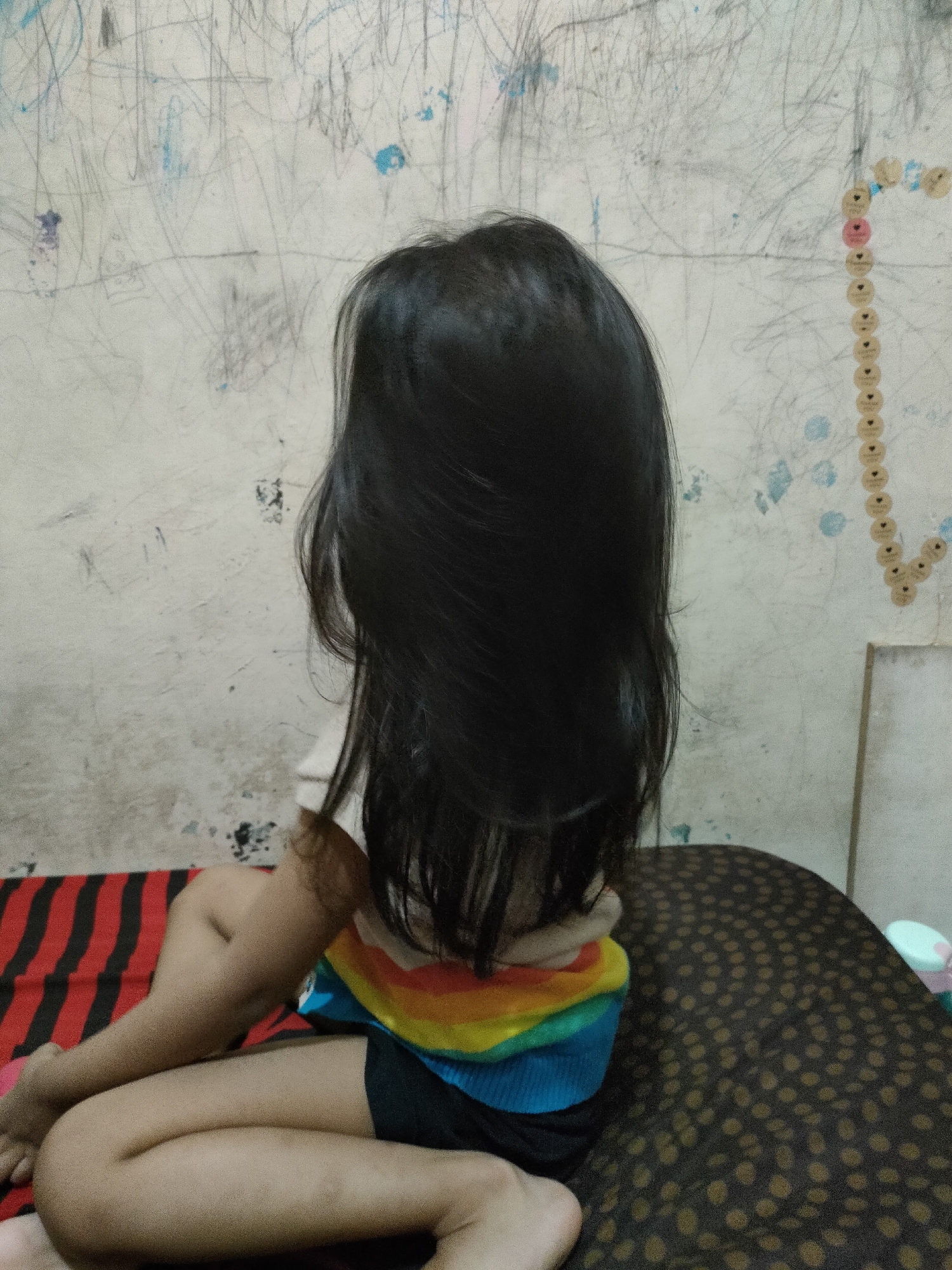 A young child with long, dark hair sits on a bed, facing away from the camera. The child wears a shirt with rainbow-colored stripes and black shorts. The bed is covered with a red and black striped blanket and a brown polka-dotted comforter. The background is a wall covered in scribbled drawings and a vertical chain of small circular stickers, some of which are heart-shaped. The scene appears to be indoors, likely in a bedroom. The location is identified as Gedangan, Indonesia.