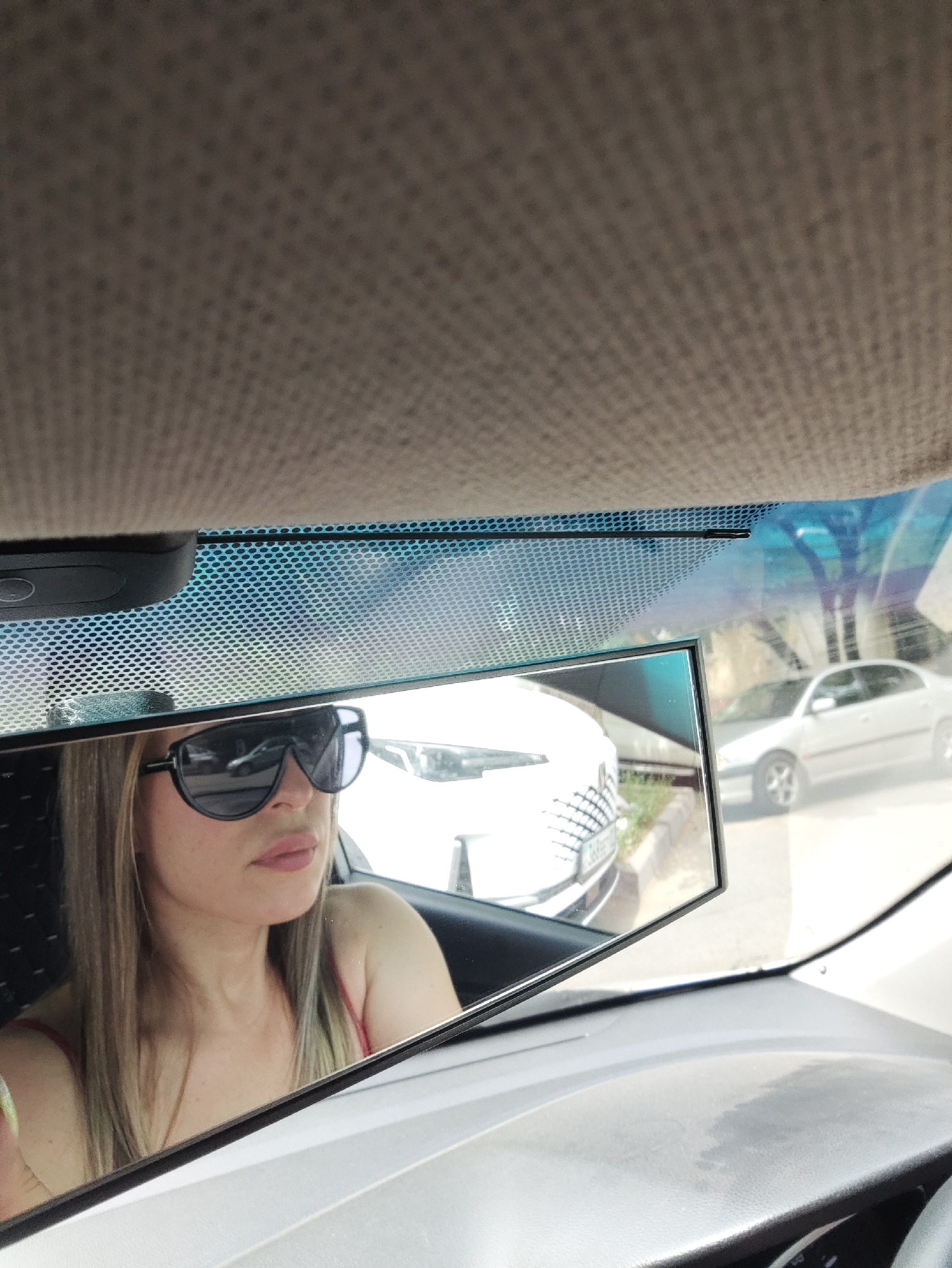 The image captures a woman wearing sunglasses and a red top, reflected in a car's rearview mirror. She is sitting in the driver's seat, with her upper body and face visible. The mirror also reflects the exterior of the car, showing another white car parked behind. The background suggests an outdoor setting with buildings and trees, likely in a city. The overall scene depicts a candid moment of a woman in a car, checking her appearance or surroundings. The location is Almaty, Kazakhstan.
