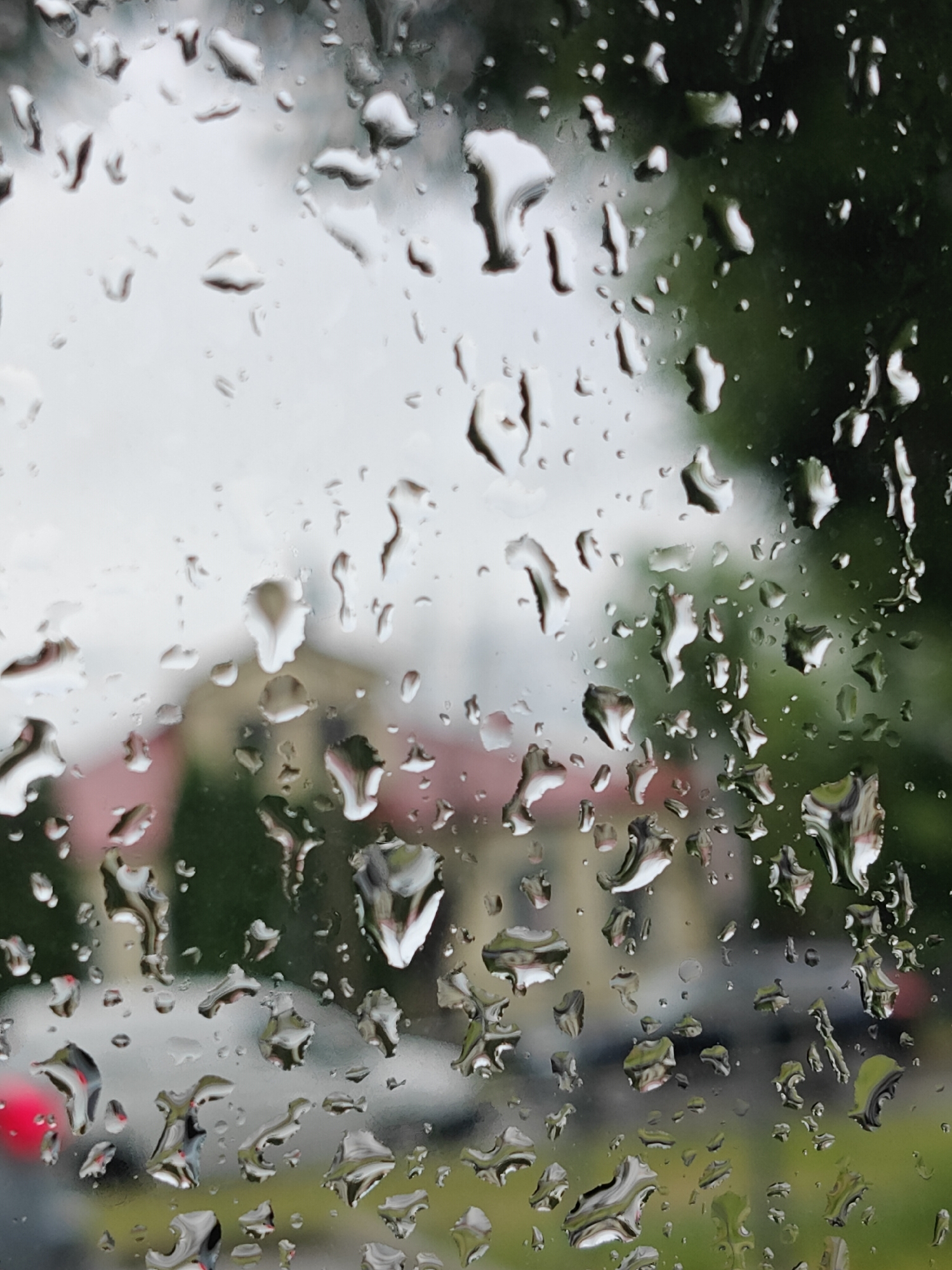 This is a close-up, medium shot of water droplets on a window pane, seen from the interior of a vehicle. The droplets distort the view of the exterior, which appears to be a city street in Pushkino, Russia. In the background, a building with a pink roof and light-colored walls is visible, along with blurred greenery, suggesting trees or bushes. The sky is overcast and appears light gray. The focus is on the water droplets, which create abstract shapes and reflections. There are no people or specific actions discernible in the image; it is a still life of a rainy day viewed through glass.