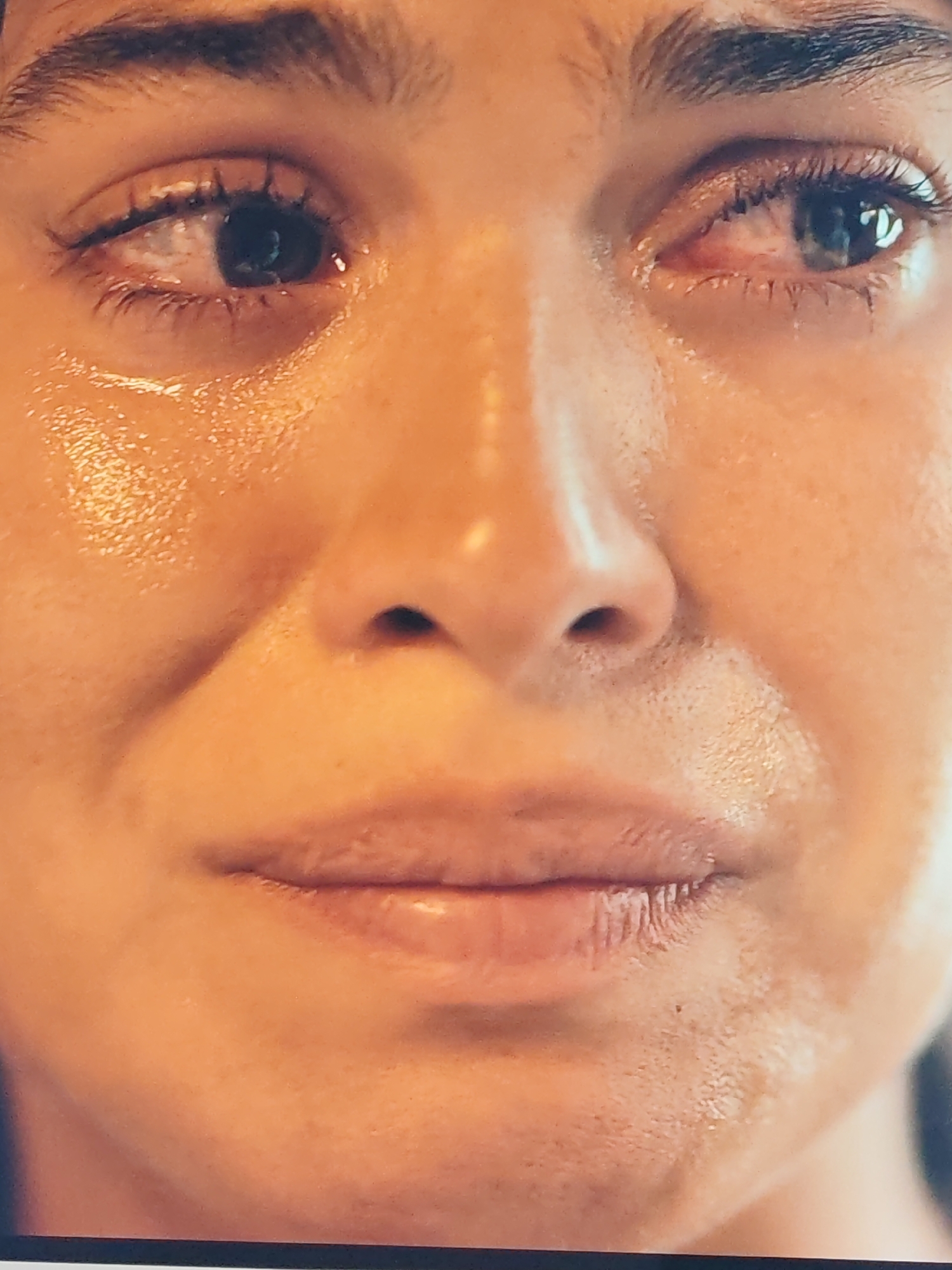 A close-up, detailed, full-frame shot of a person's face, with a focus on the eyes, nose, and mouth. The subject is crying, with tears visible on the cheeks and around the eyes. The skin appears slightly glossy due to moisture. The lighting is warm and highlights the texture of the skin. The background is blurred, suggesting an indoor setting or a shallow depth of field. The image does not provide any information about the location within a city or any specific environment.