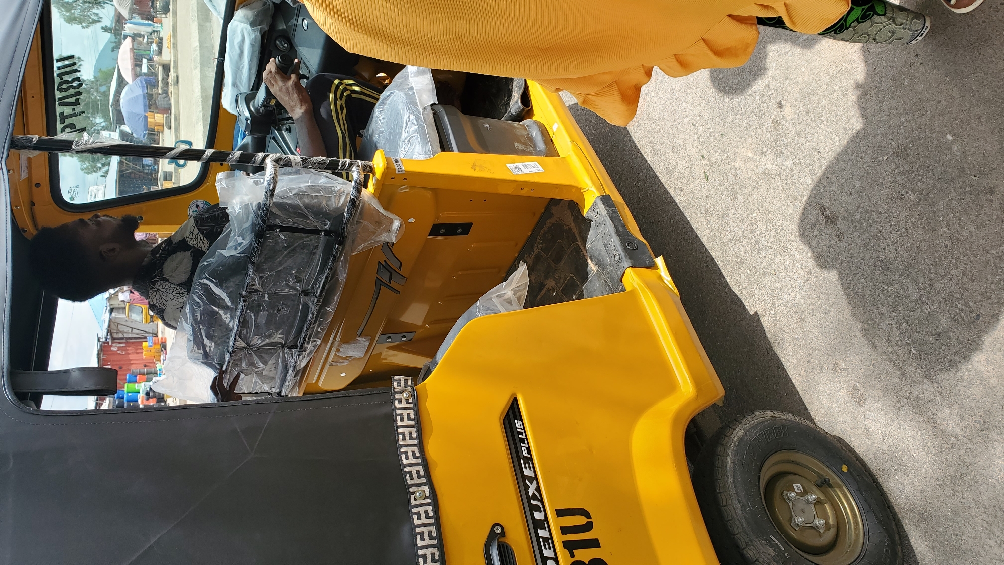 In Jalingo, Nigeria, a yellow three-wheeled auto-rickshaw, identified with "DELUXE PLUS 81U" branding, is parked on a paved surface. The driver, a Black man with short, dark, afro-style hair and a beard, is seated in the driver's seat, looking to his right. He is wearing a black and white patterned short-sleeved shirt and dark pants with yellow stripes. His right hand is on the steering wheel. The interior of the auto-rickshaw is partially covered in clear plastic sheeting. A person wearing a bright orange garment is partially visible on the right side of the image, standing outside the auto-rickshaw. The background shows a street scene with various stalls, umbrellas, and buildings, suggesting a market or commercial area. Sunlight casts shadows on the ground.