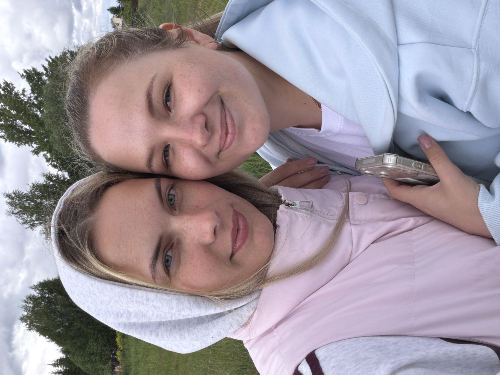 Two young women are posing closely for a selfie outdoors. One woman is wearing a light blue hooded sweatshirt, and she is holding a cell phone with a clear case. The other woman is wearing a pink and light grey hooded vest. The background consists of trees, grass, and an overcast sky. The location is Ольховка, Russia.
