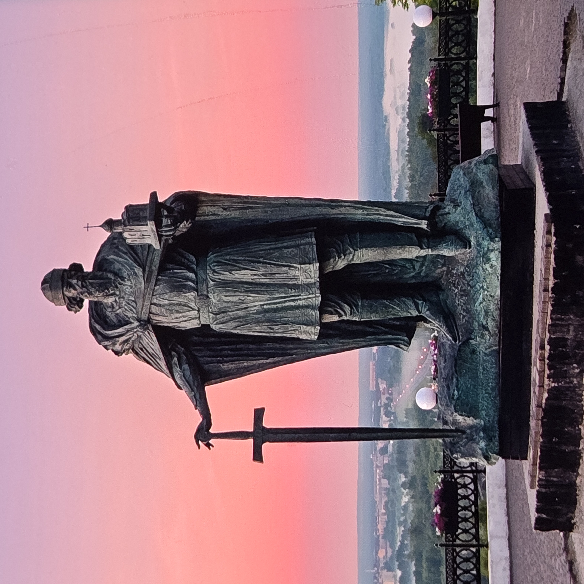 The image shows a full-body bronze statue of a historical figure, possibly Vladimir the Great, standing on a stone pedestal. The statue depicts a bearded man wearing a helmet, a long cloak, and a tunic, holding a sword in his left hand and a model of a church in his right. The statue is positioned outdoors, overlooking a city with buildings and greenery in the background, shrouded in a light mist. The sky behind the statue is a gradient of pink and purple, indicating either sunrise or sunset. The statue is located in the city of Vladimir, Russia.