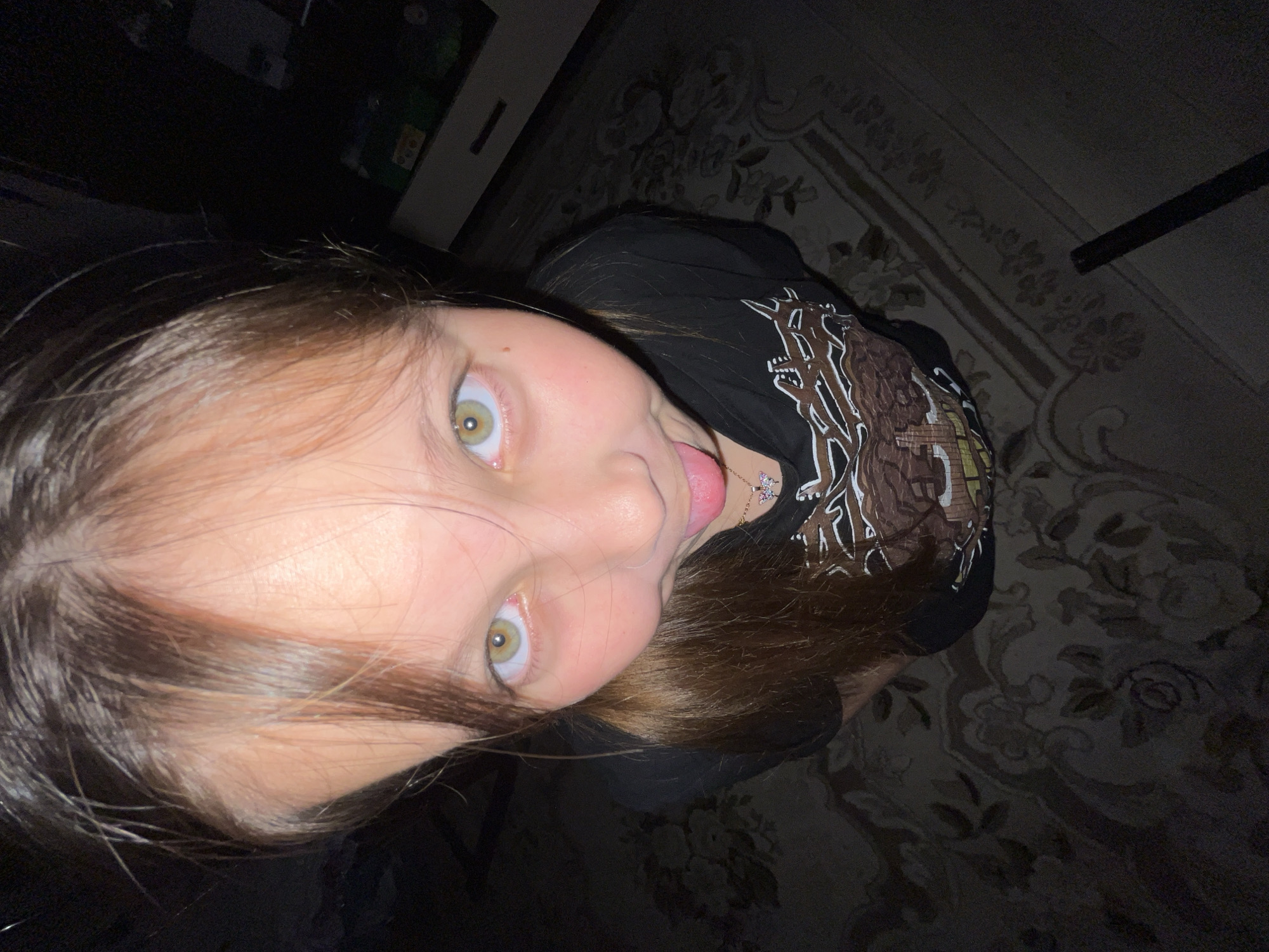 A young girl with light skin and light brown hair is looking up at the camera with a playful expression, sticking her tongue out slightly. Her eyes are light green. She is wearing a black t-shirt with a brown and white graphic design. A small necklace with a butterfly pendant is visible. The background includes a patterned rug and a dark area that appears to be a room interior. There is no verifiable information to suggest the location is in the city of Novopokrovskaya, Russia.