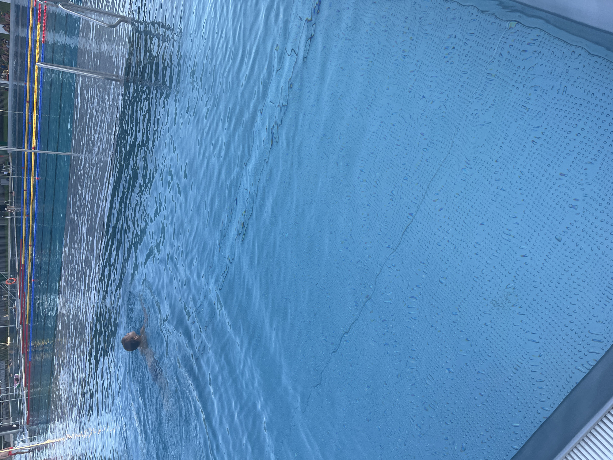 A child is swimming in a blue swimming pool. The child is in the upper left quadrant of the image, facing away from the camera. The child is wearing dark swim trunks and has short brown hair. The water is clear and blue, with ripples on the surface. The pool appears to be outdoors, with lanes marked by buoys and ropes. In the background, there are people sitting on the edge of the pool and a lifeguard stand. The pool has a tiled bottom. The image is taken from a high angle, looking down into the pool.