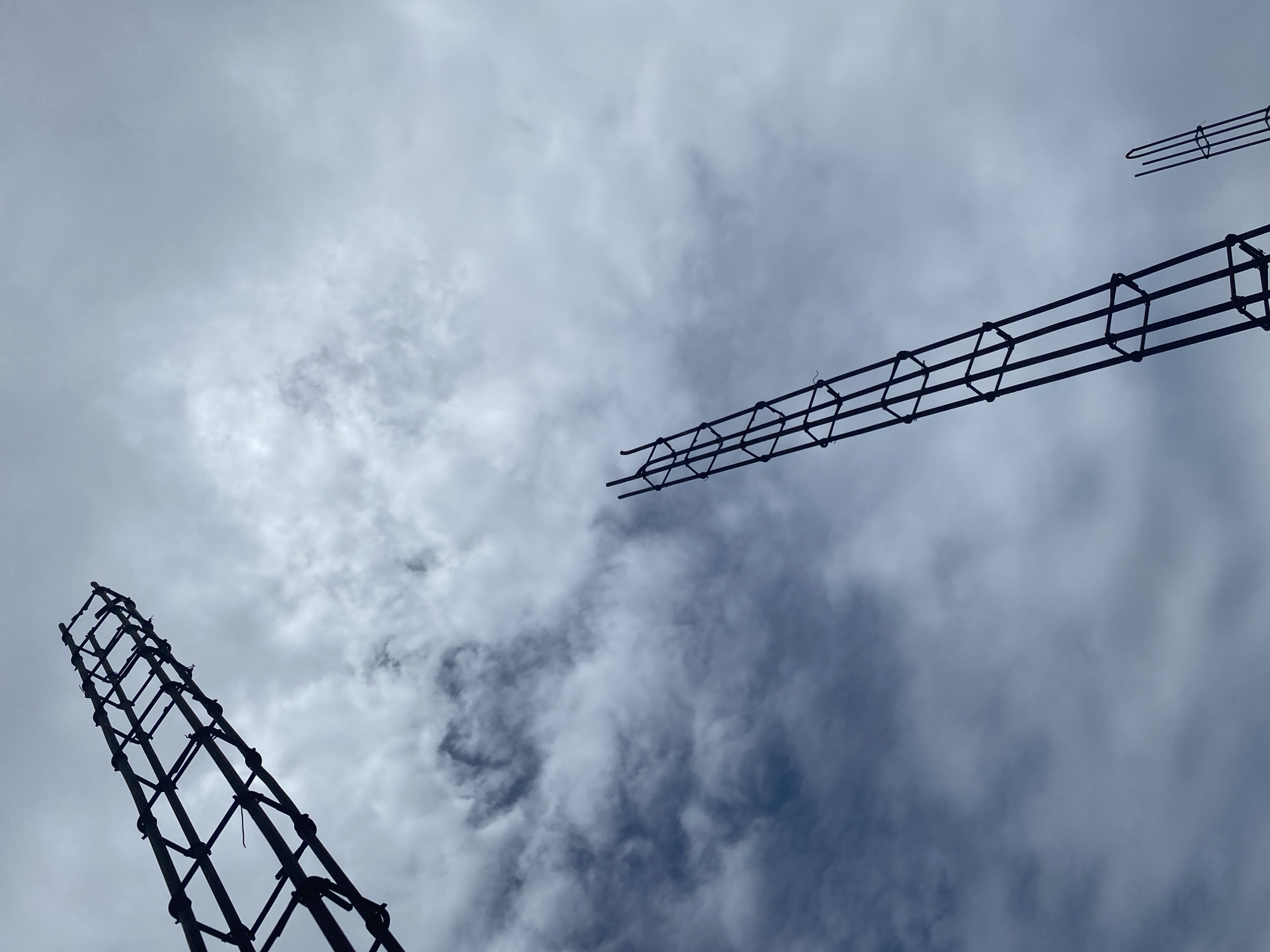 In an outdoor scene, three dark, skeletal rebar columns pierce a cloudy, overcast sky. The columns, constructed from vertical and horizontal steel bars, form rectangular grids and are viewed from below, looking upwards. The rebar is dark against the lighter, textured clouds. The environment is anonymous, lacking any discernible urban or natural landmarks. No individuals are present. The scene suggests a construction site or an unfinished structure, with the rebar acting as foundational support for future building elements.