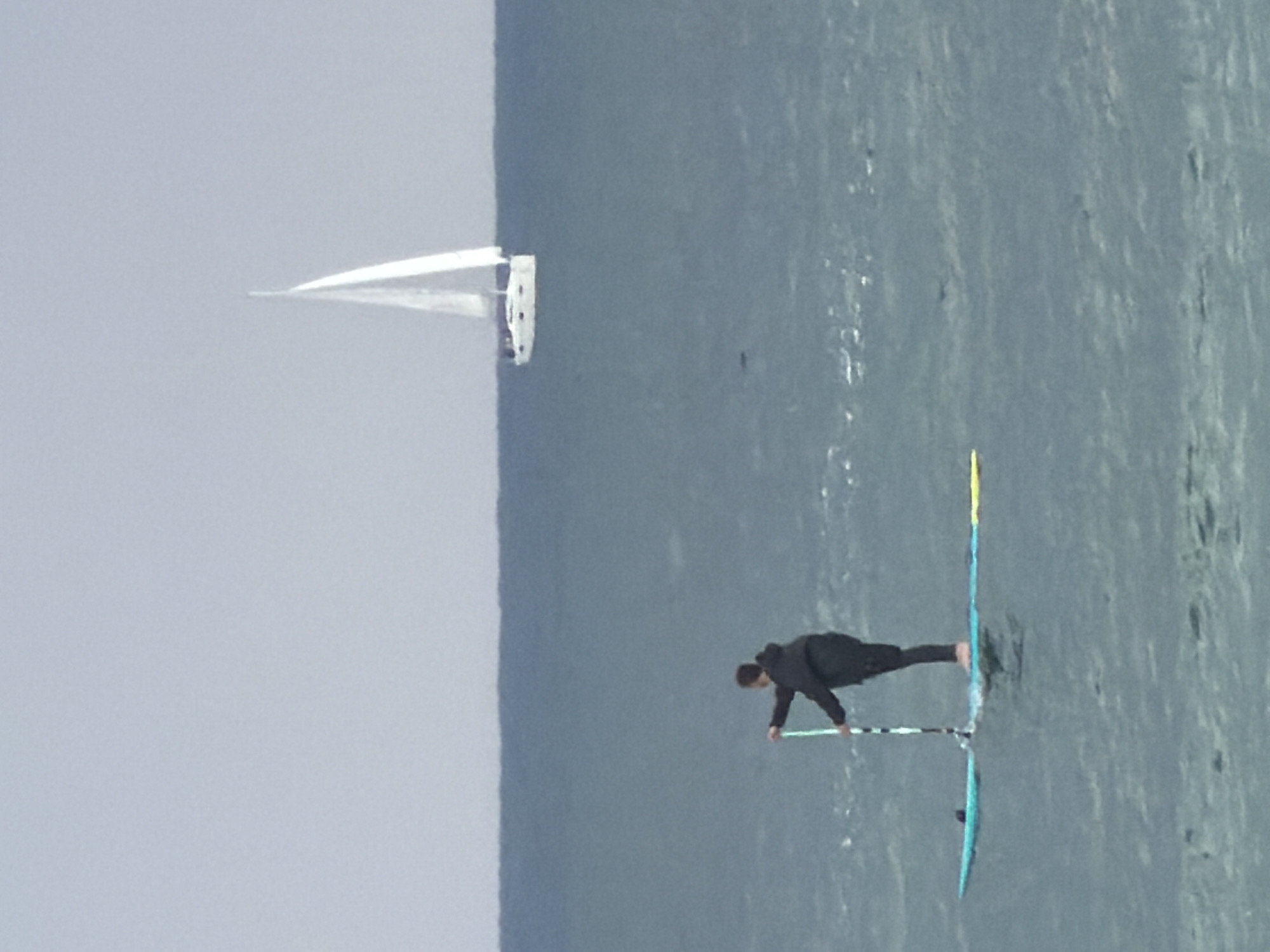 A landscape view depicts a body of water under a cloudy sky. On the water, a windsurfer, clad in black, is actively engaged in windsurfing. Separately, a sailboat is seen in the distance. The scene likely takes place off the coast of Tel Aviv, Israel.