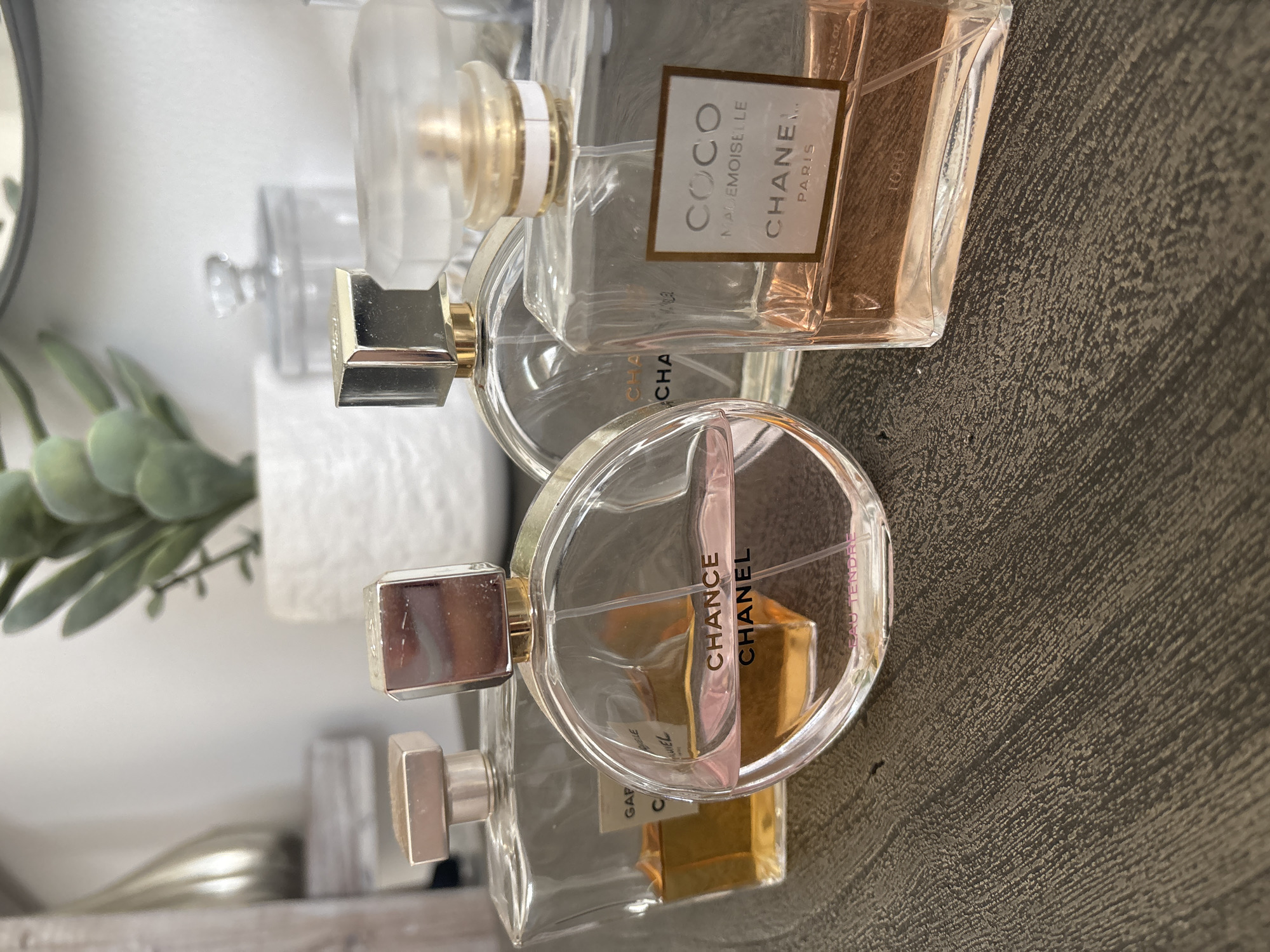 Four Chanel perfumes are displayed on a dark wooden surface. From left to right, the perfumes are: Chance Eau Tendre, Chance, Coco Mademoiselle, and Gabrielle. The background is blurred and shows a white wall, a white pot with a green plant, and a reflective surface. The lighting is soft and natural. The scene appears to be indoors. There is no indication of the location being in Dubai, United Arab Emirates.