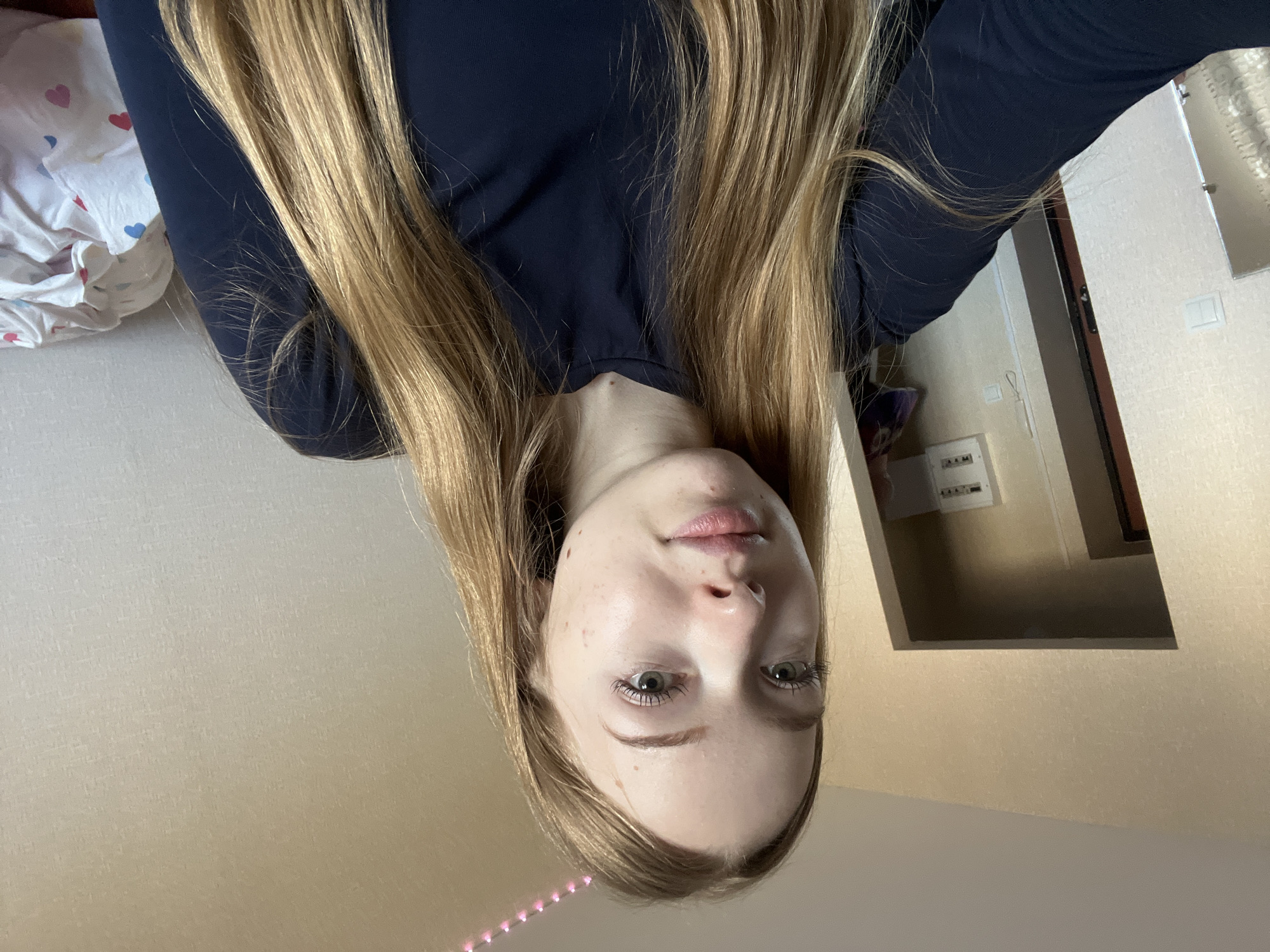 The image shows a young woman with long blonde hair, wearing a dark blue shirt. Her face is visible, showing light skin, green eyes, and some minor skin blemishes. The background includes a bed with patterned bedding, a wall with an electrical box, a partially visible doorway and a section of LED light strip. The orientation of the image suggests it was taken at an unusual angle, with the subject positioned upside-down in relation to the environment. There is no information to indicate the location is specifically in Chaikovsky, Russia.