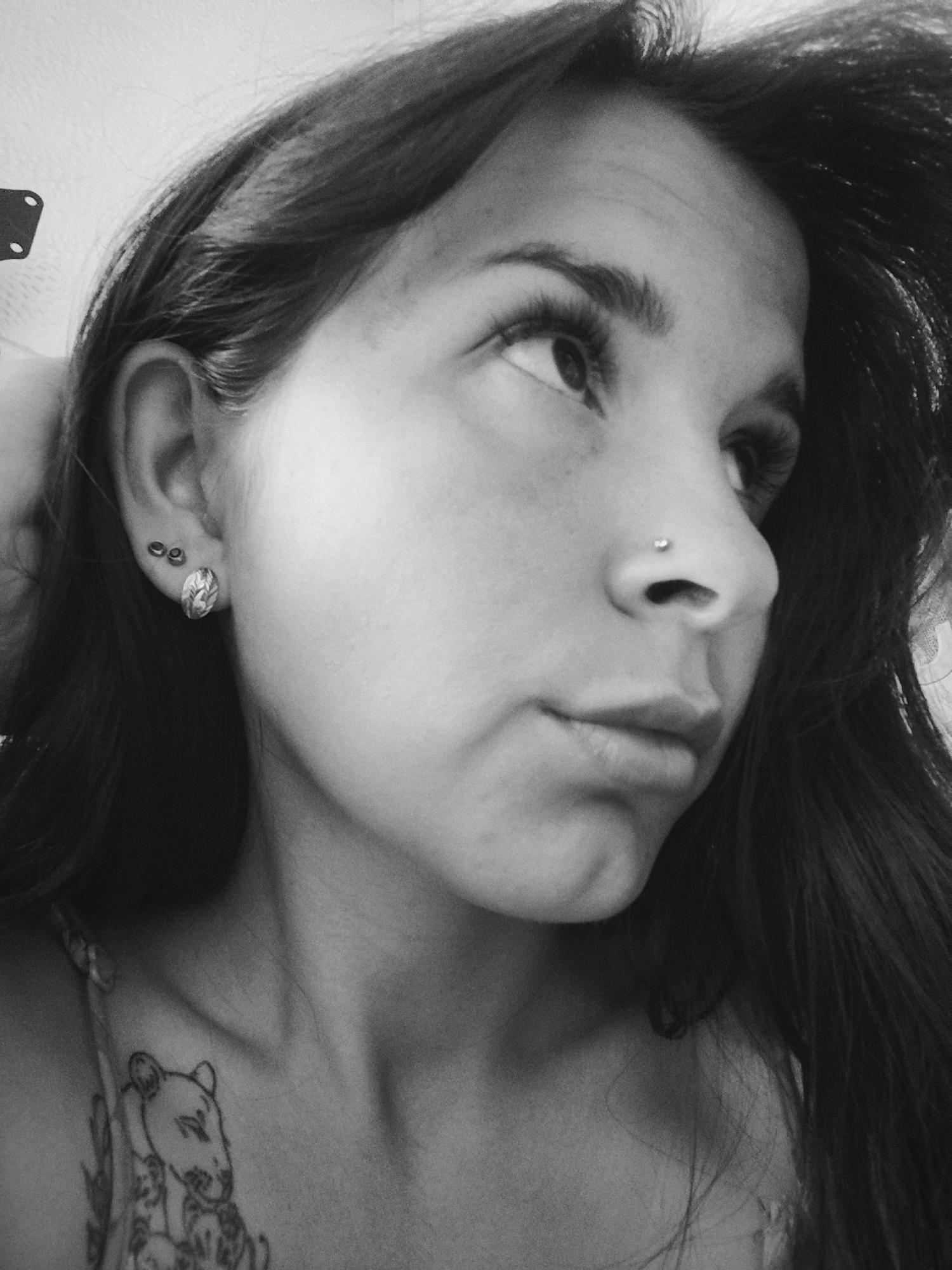 A black and white medium shot depicts a person with long dark hair looking upwards and to the right. Visible piercings include a nose stud and two earrings in the left ear (one hoop and one larger stud). A tattoo of a lioness is on the person’s chest. The location is not discernible.