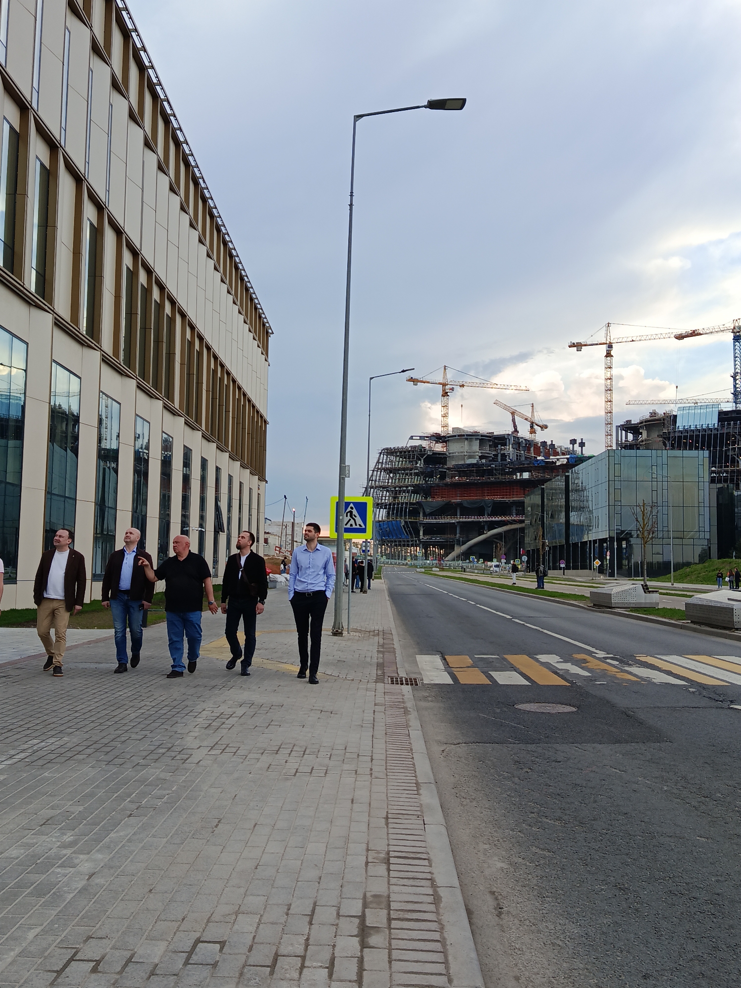 The image depicts a street scene in Moscow, Russia. Five men are walking on a paved sidewalk along the left side of the frame. To their left is a modern, multi-story building with numerous rectangular windows. In the background across the street are ongoing construction projects, indicated by cranes, scaffolding, and partially completed structures. A pedestrian crosswalk is visible on the road, with yellow and white stripes. The sky is overcast, with a mix of light and dark clouds. Streetlights and road signs are positioned along the street.