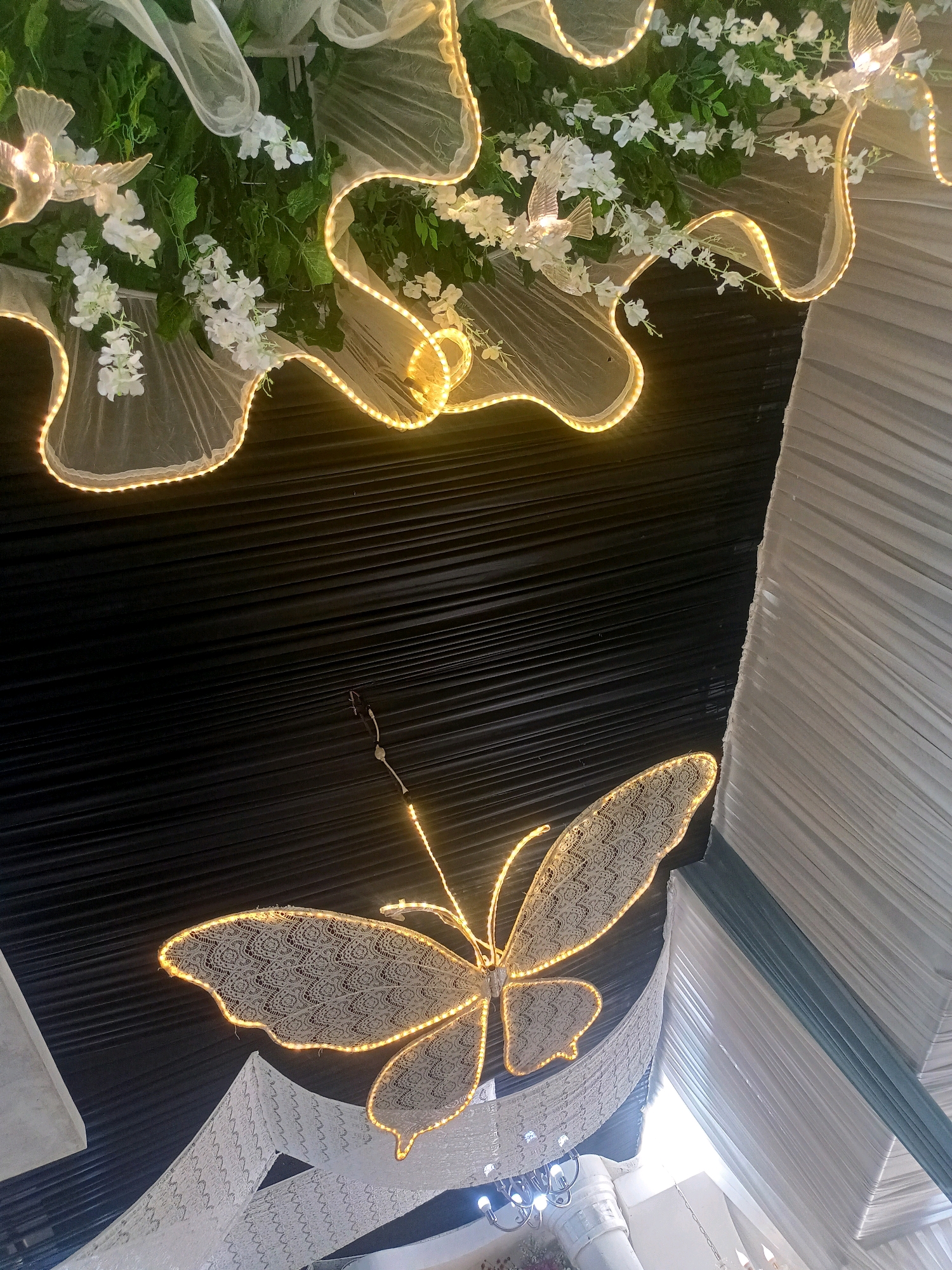 The image displays a decorative ceiling installation, likely at an event venue in Badamita, Indonesia. A large, illuminated butterfly made of white lace and outlined with warm-toned LED lights is the central focus. It hangs suspended against a dark, textured background. Above the butterfly, a white, sheer fabric draped with more LED lights and artificial white flowers and decorative birds is visible. White pleated fabric drapes line the sides of the frame, suggesting a tent-like or banquet hall setting. The scene is illuminated by the warm glow of the lights. No people are visible in the image.