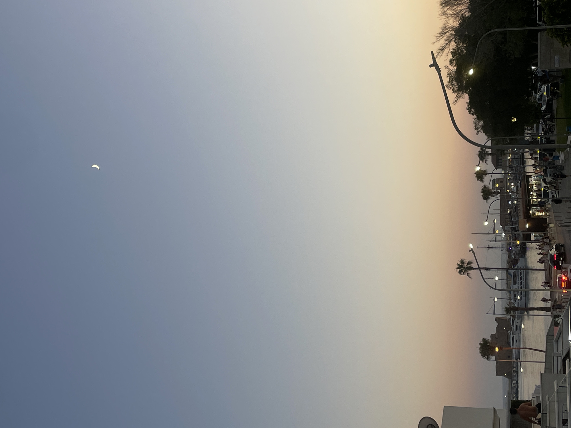 The image depicts a scenic twilight view of the Paphos Harbour in Cyprus. The sky transitions from a clear pale blue at the top to soft shades of orange and pink near the horizon. A crescent moon is visible in the upper portion of the sky.
In the foreground, a man is partially visible on a white structure. To the right, a lamppost with a modern design curves over a dark, leafy tree. Further into the scene, the harbour is filled with various boats, including yachts and smaller vessels. Across the water, a historic stone structure, likely a castle, stands prominently.
Along the waterfront, there is a pedestrian walkway with several streetlights illuminating the area. People are seen walking and gathered in small groups. Cars are visible on the road, with their headlights on, suggesting a transition into the evening. Palm trees are scattered throughout the scene, adding to the Mediterranean ambiance. The overall impression is that of a tranquil evening at a bustling harbour.
