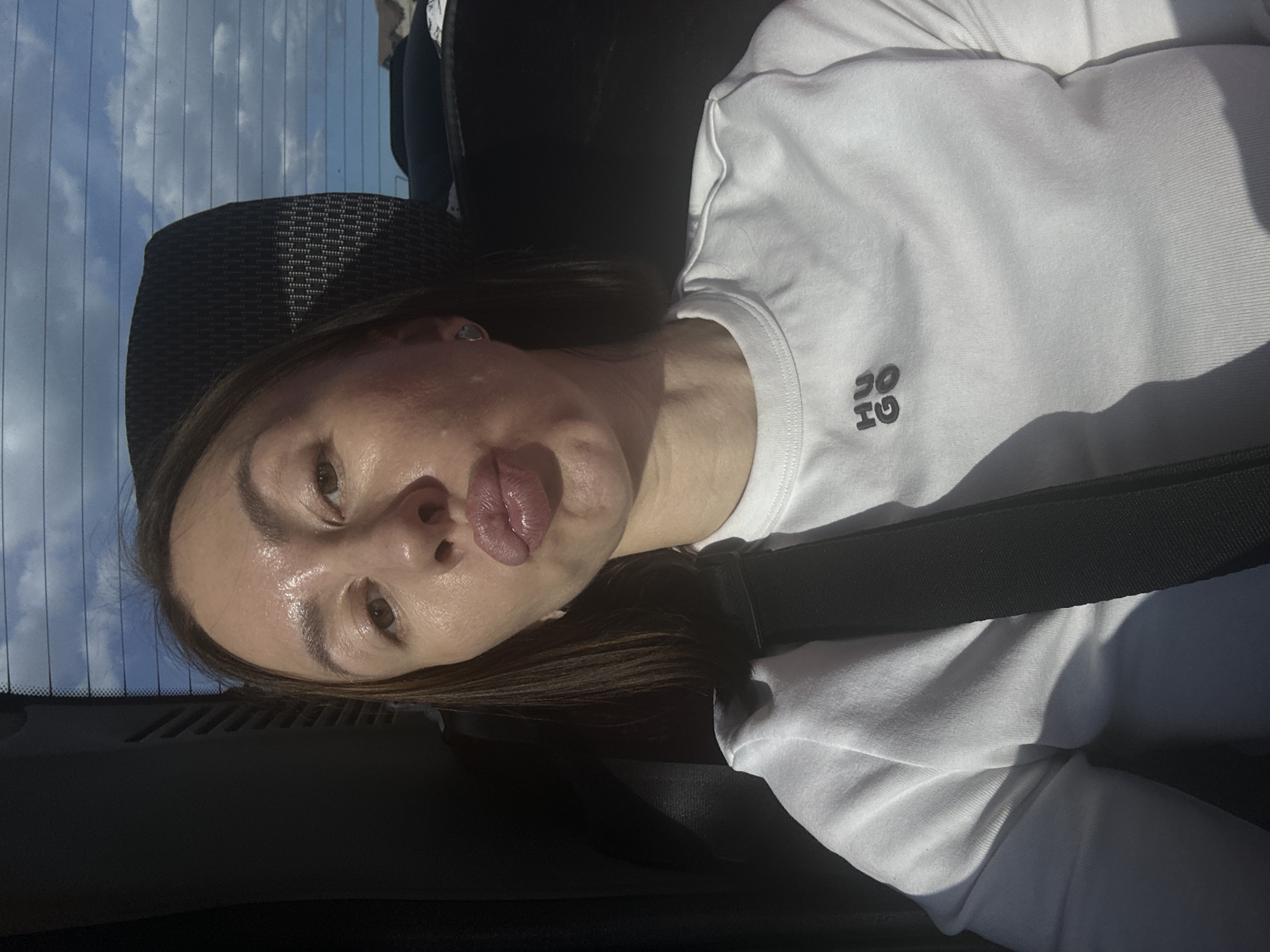 A selfie of a woman with brown hair wearing a white long-sleeved shirt with the Hugo Boss logo on the shoulder. The woman is in the backseat of a car wearing a seatbelt. The car appears to be in the city of Bolgury, Russia, as indicated by the sky visible through the car window.