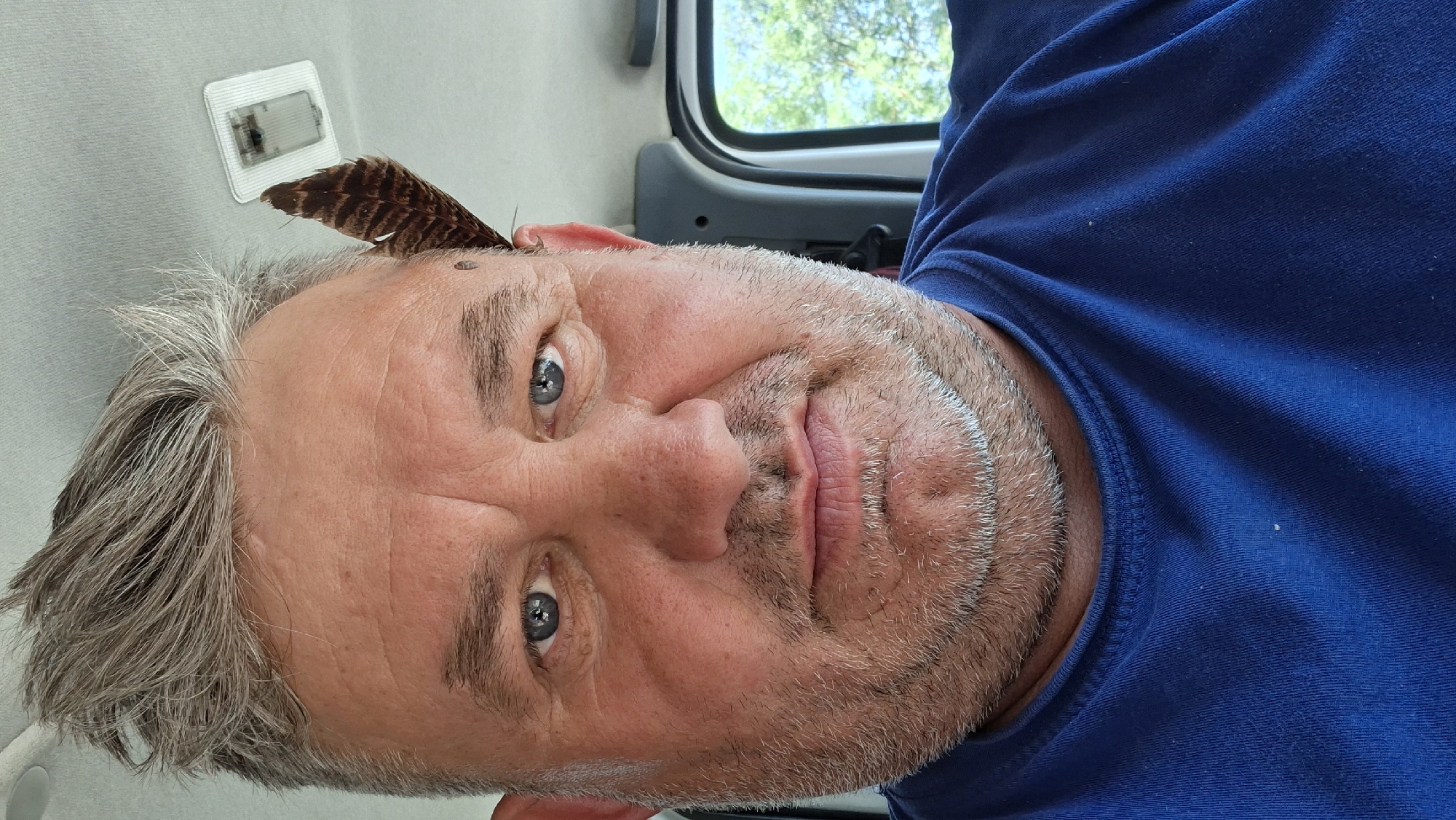 A close-up, selfie-style photograph shows a middle-aged man with graying hair, blue eyes, and a light beard. He is wearing a dark blue t-shirt. A brown and black feather is lodged in his hair near his right temple. The man appears to be inside a vehicle, as indicated by the interior light fixture visible on the ceiling and the blurred greenery seen through a window on the right side of the frame. The background suggests an outdoor environment, possibly a park or a tree-lined road, common in areas like Hîncești, Moldova, which is characterized by its numerous green spaces and rural outskirts. The lighting is natural, coming from the outdoor environment.