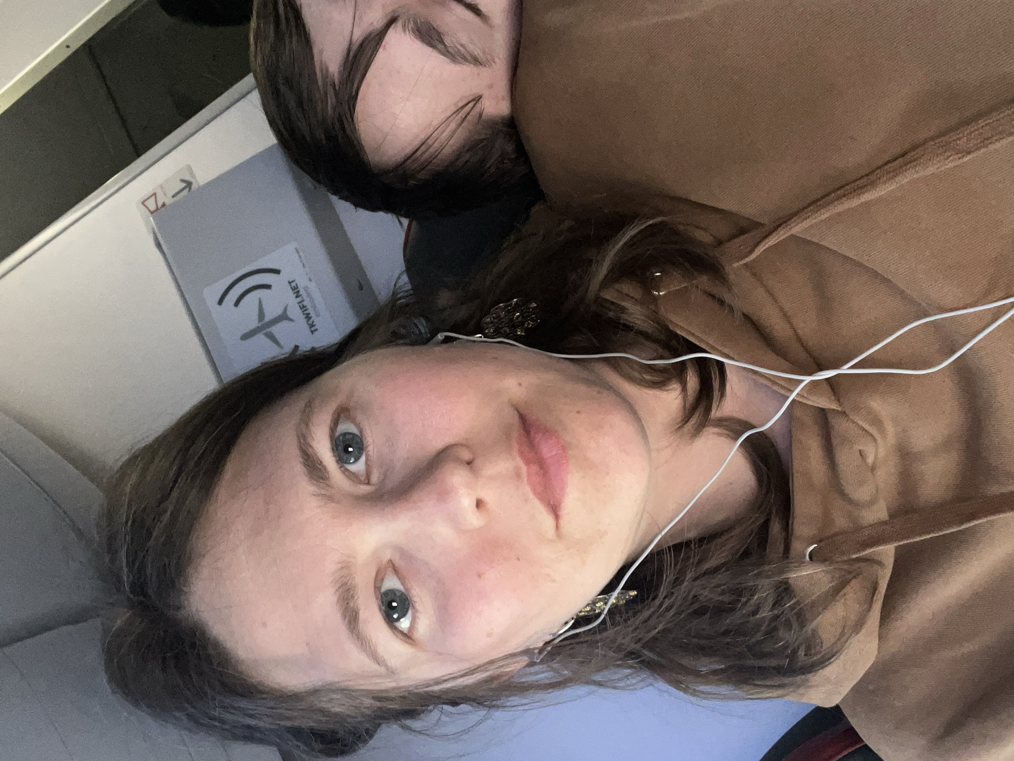 A close-up portrait, taken inside an aircraft cabin, features two individuals. In the foreground, a woman with brown hair and blue eyes looks directly at the camera with a neutral expression. She is wearing a brown hooded sweatshirt and white wired earphones are plugged into her ears. An ornate, dark-colored earring is visible on her right earlobe.
Behind and slightly to the right of the woman, a second person, likely a companion, is partially visible, with dark hair and their forehead resting, suggesting they are asleep or resting their head.
To the left of the woman, a gray airplane panel or compartment is visible. Adhered to this panel is a white sticker with "TK WIFI.NET" printed on it, accompanied by a Wi-Fi symbol and a stylized airplane icon, indicating in-flight internet service, likely from Turkish Airlines (TK is the IATA code). Above this, a smaller sticker with a red border displays an arrow pointing right and a character resembling a 'Z' or '7'. The overall setting suggests travel, with the context indicating a flight associated with Podgorica, Montenegro. The lighting is artificial, typical of an airplane interior.