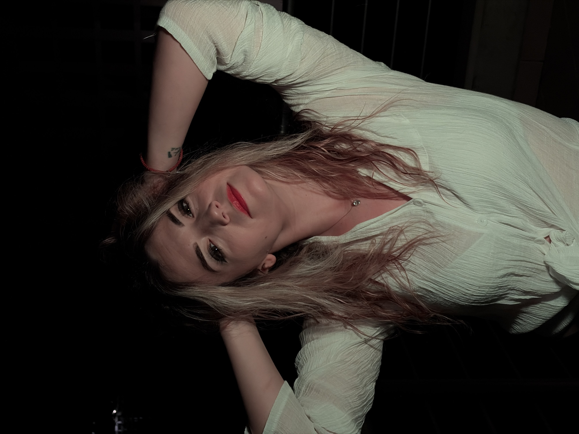 A female subject is depicted from the chest up against a predominantly black background in the city of Budva, Montenegro. She has long, light-colored hair, styled with both hands placed behind her head. Her face, angled slightly to the right and looking forward, is directly illuminated, highlighting red lipstick and dark eye makeup. She is wearing a white, textured, long-sleeved shirt with a collared V-neck, tied in a knot at the front, and an undergarment is faintly visible beneath the sheer fabric. A silver pendant necklace is worn around her neck, and a red bracelet is visible on her left wrist, along with a small tattoo on her left upper arm. The background is uniformly dark, with only indistinct, extremely faint lights discernible in the upper left corner, consistent with an outdoor nighttime environment.